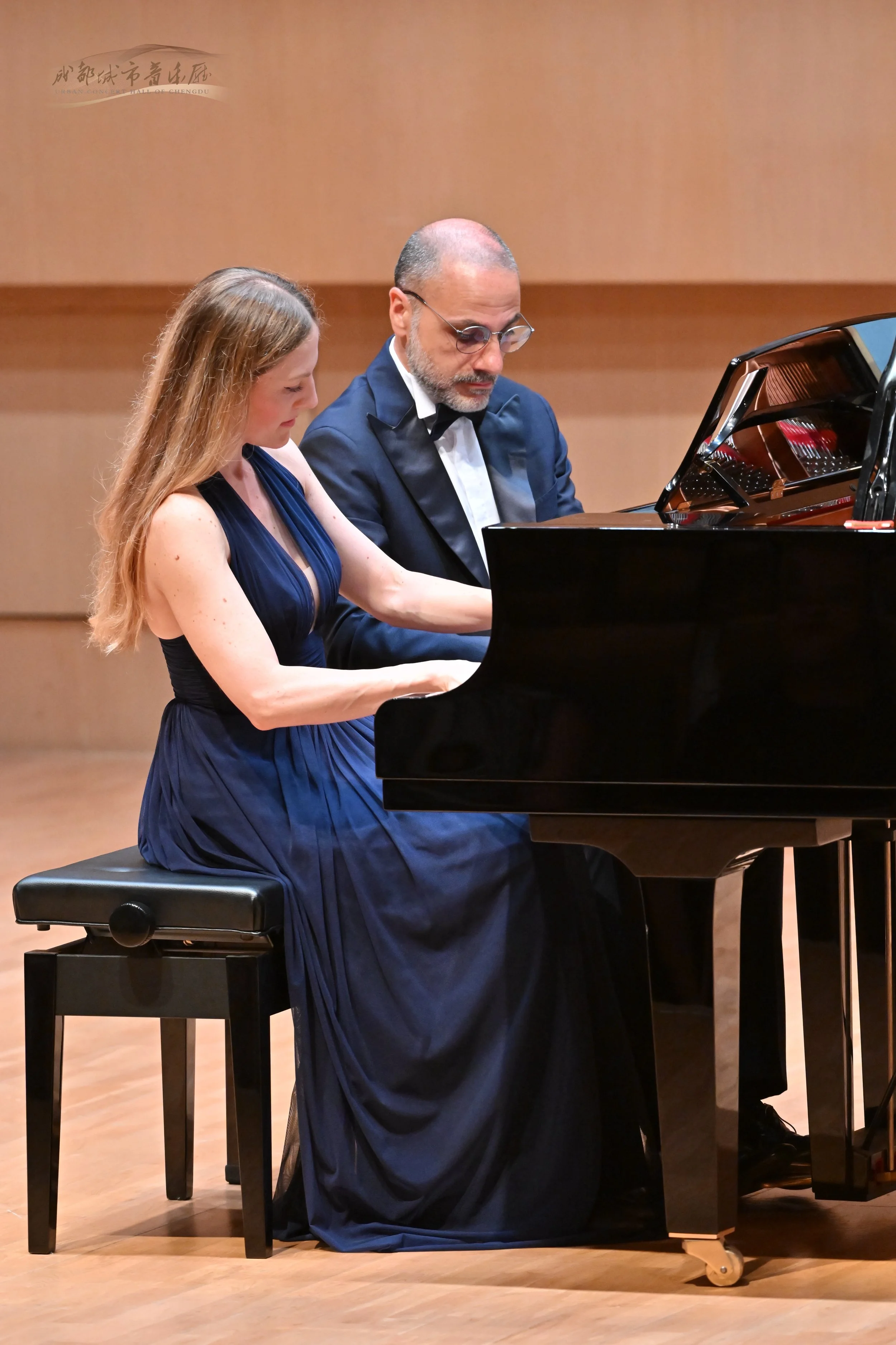 Una donna in abito blu scuro e un uomo in frac suonano un pianoforte in un concerto.