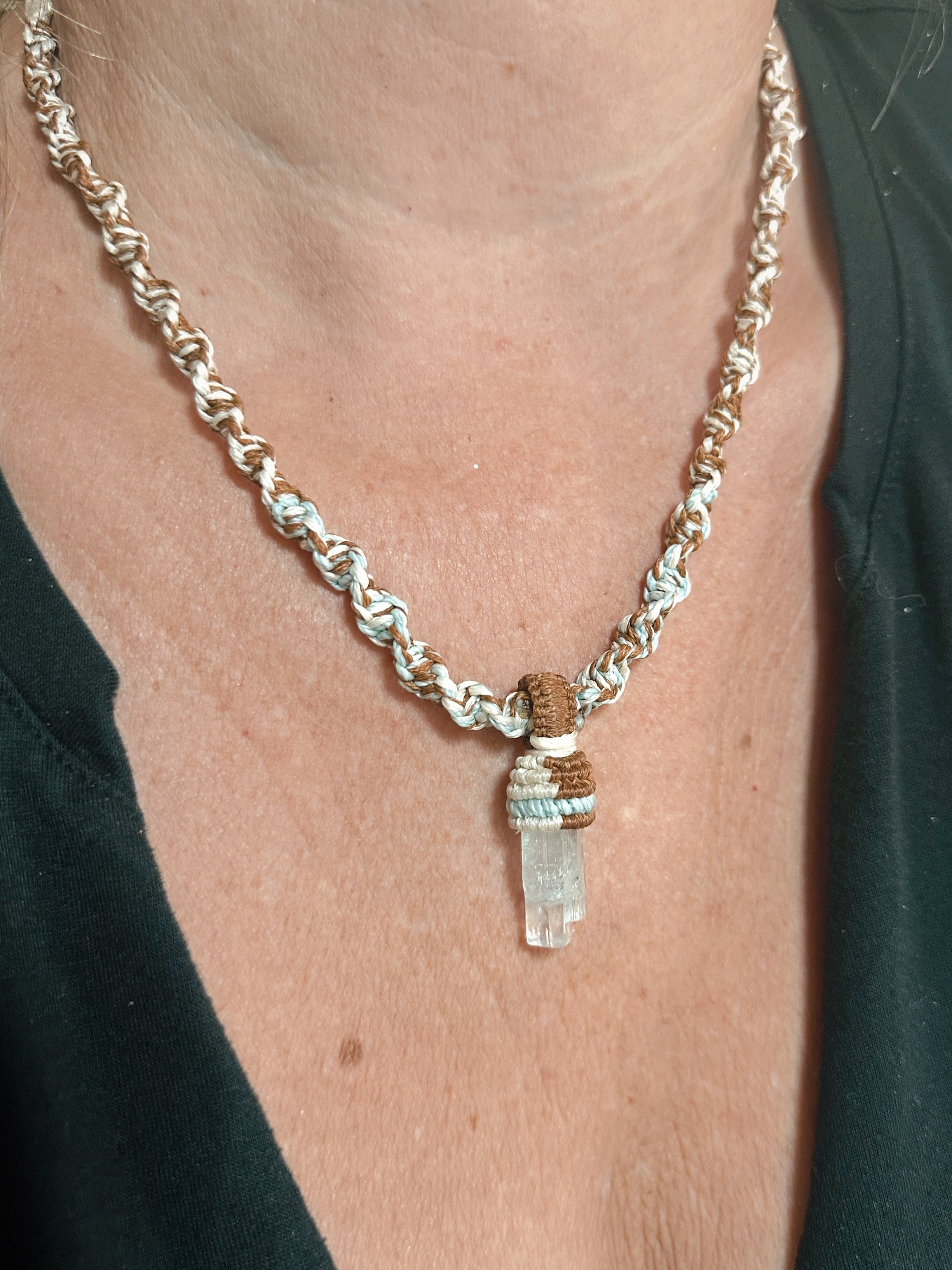 Close-up of a woman's neck and chest showing a necklace with a clear crystal pendant and colorful woven beads, with a black top and green outerwear.