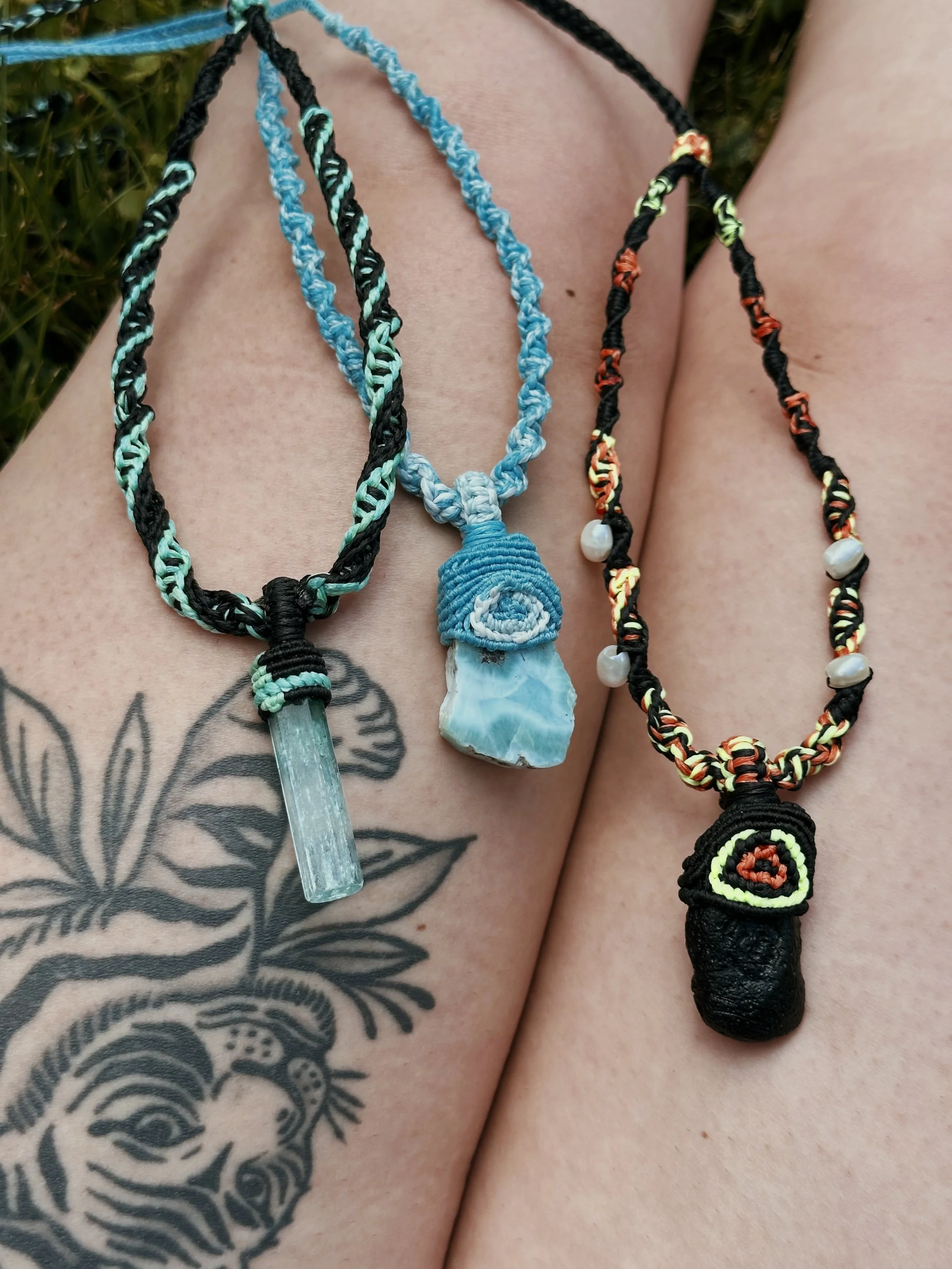 Three handmade friendship necklaces with colorful braided cords and pendants: one with a pointed crystal, one with a spiral shell, and one with a small fabric-covered pendant with a spiral pattern, all resting on a person's tattooed chest.
