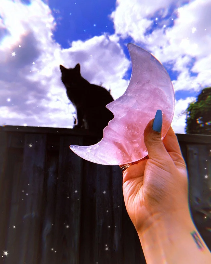 A person's hand holding a pink moon-shaped crystal against a vibrant blue sky with white clouds and star-like sparkles; a black cat sits on a fence in the background.