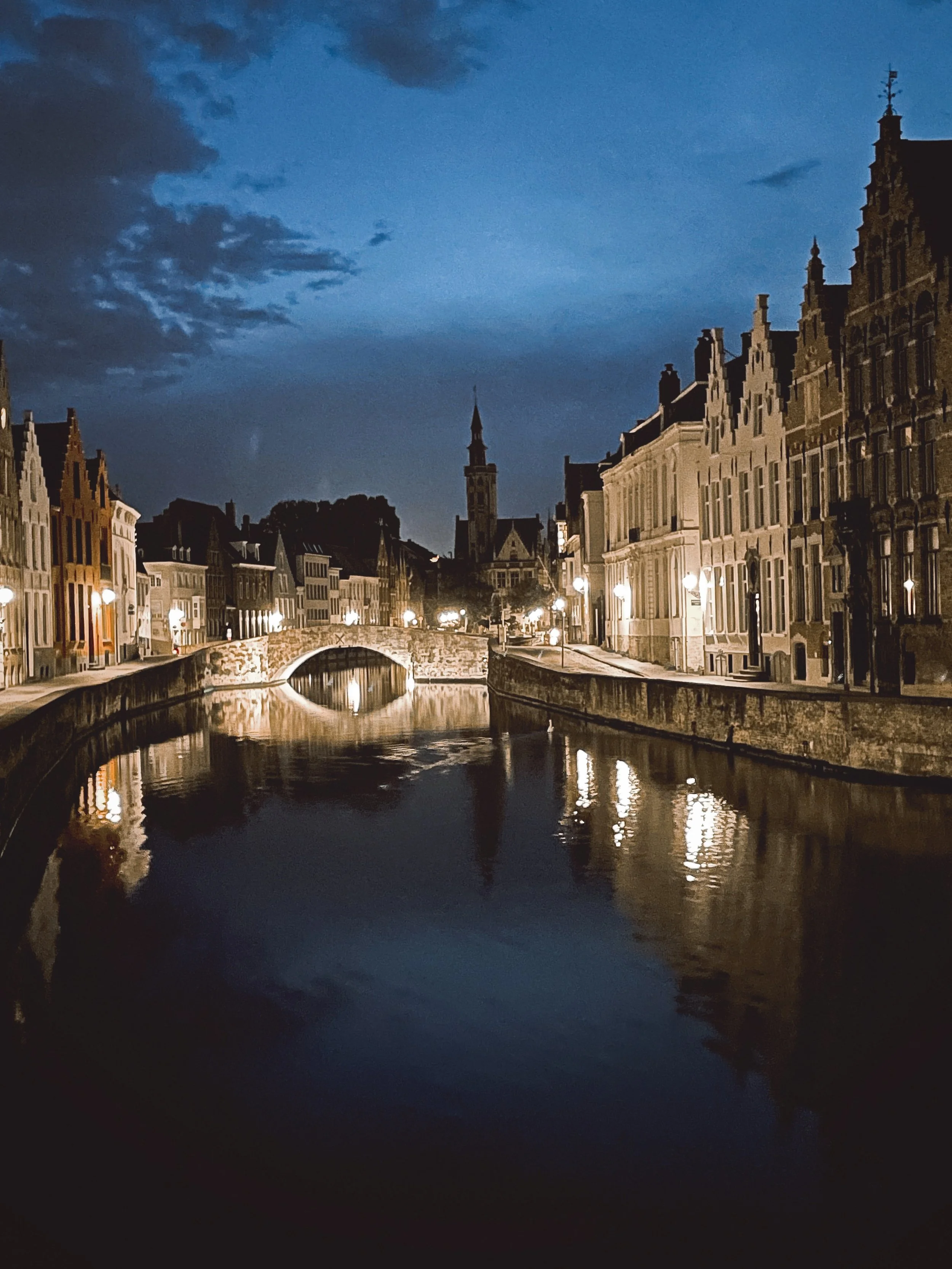 A nighttime view of a European city with a canal, historic buildings, and a bridge, illuminated by streetlights, with a cloudy sky overhead.