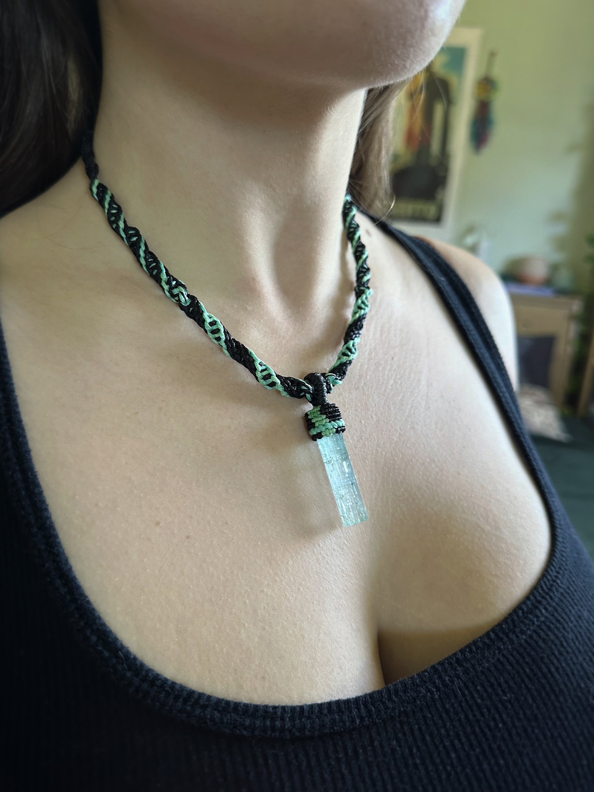 Close-up of a person's neck and chest, wearing a black top with a woven necklace that has a light blue pendant, and a blurred background with various objects.