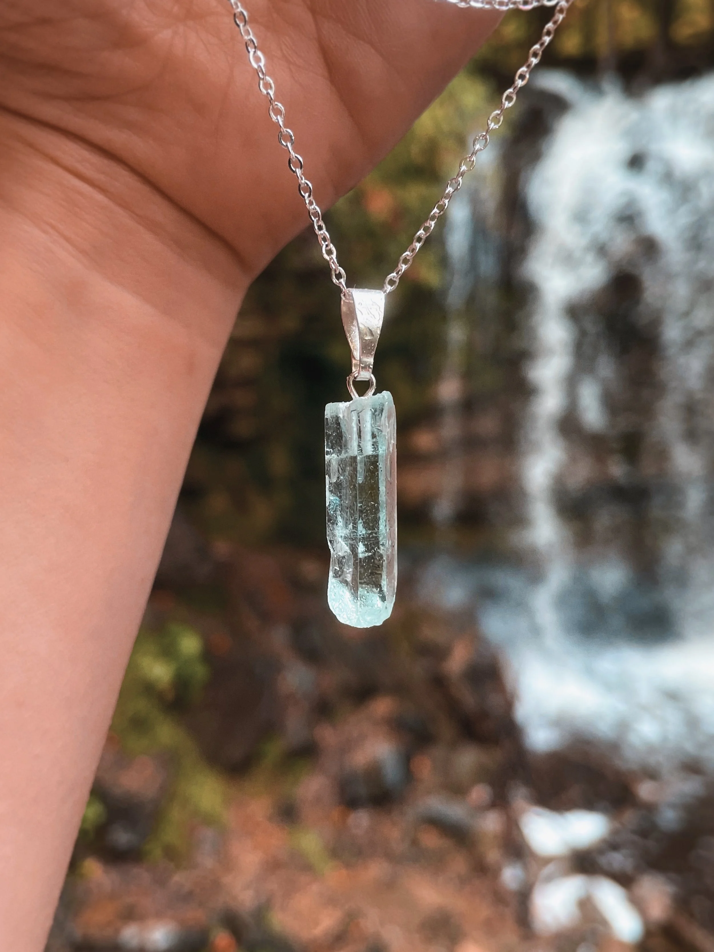 Close-up of a silver necklace with a rectangular crystal pendant hanging from a chain, held against a blurred outdoor background with rocks and water.