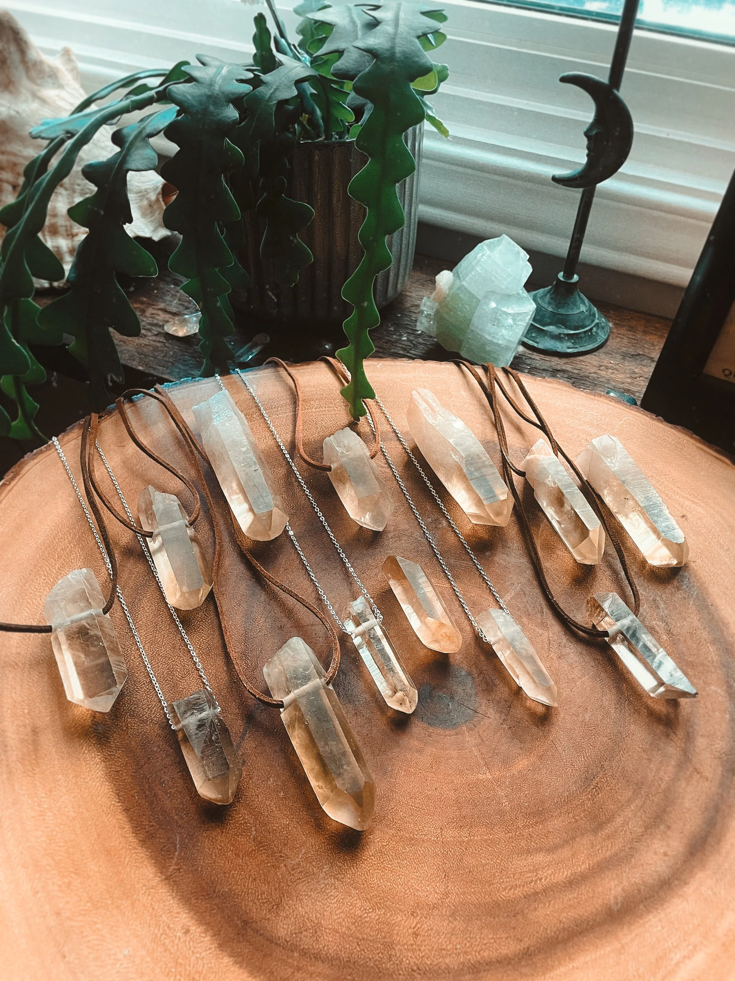 A display of various crystal necklaces on a leather surface, with a plant, a crystal, and a moon-shaped candle holder nearby, all arranged in front of a window with blinds.