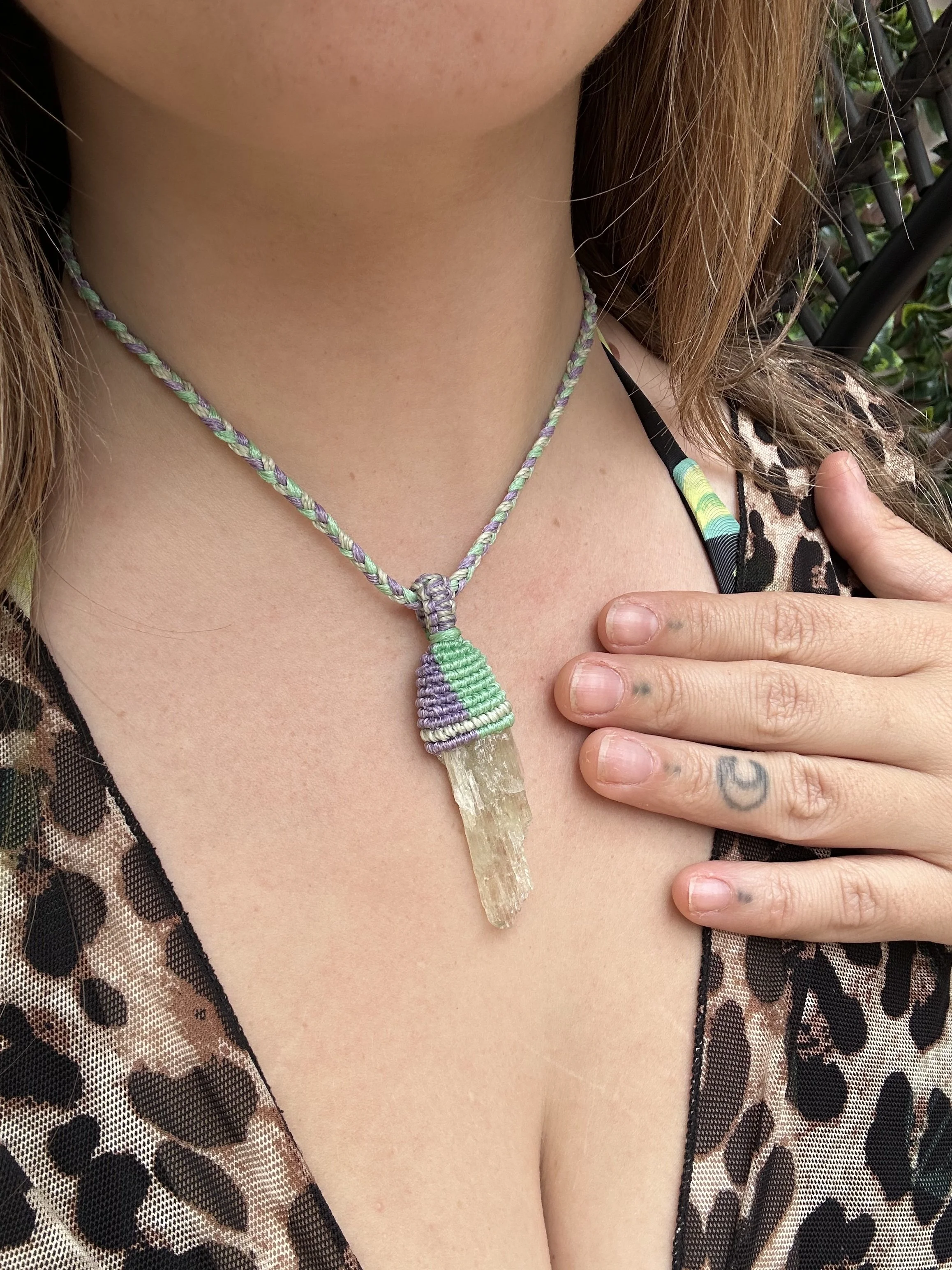 Close-up of a woman wearing a multi-colored woven necklace with a large, transparent crystal pendant, and a leopard print garment. Her hand, which has small tattoos, rests on her chest.