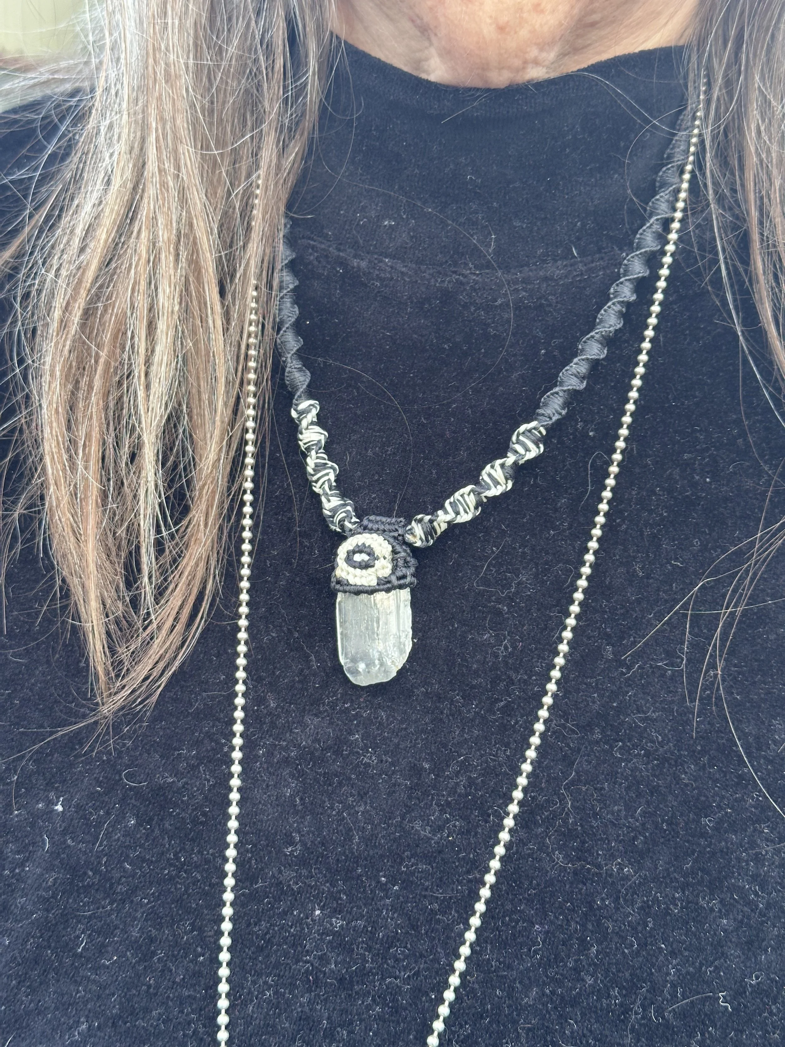Close-up of a person wearing layered necklaces, including a beaded white necklace, a black and white patterned necklace, and a pendant with a gray crystal or stone on a black background.