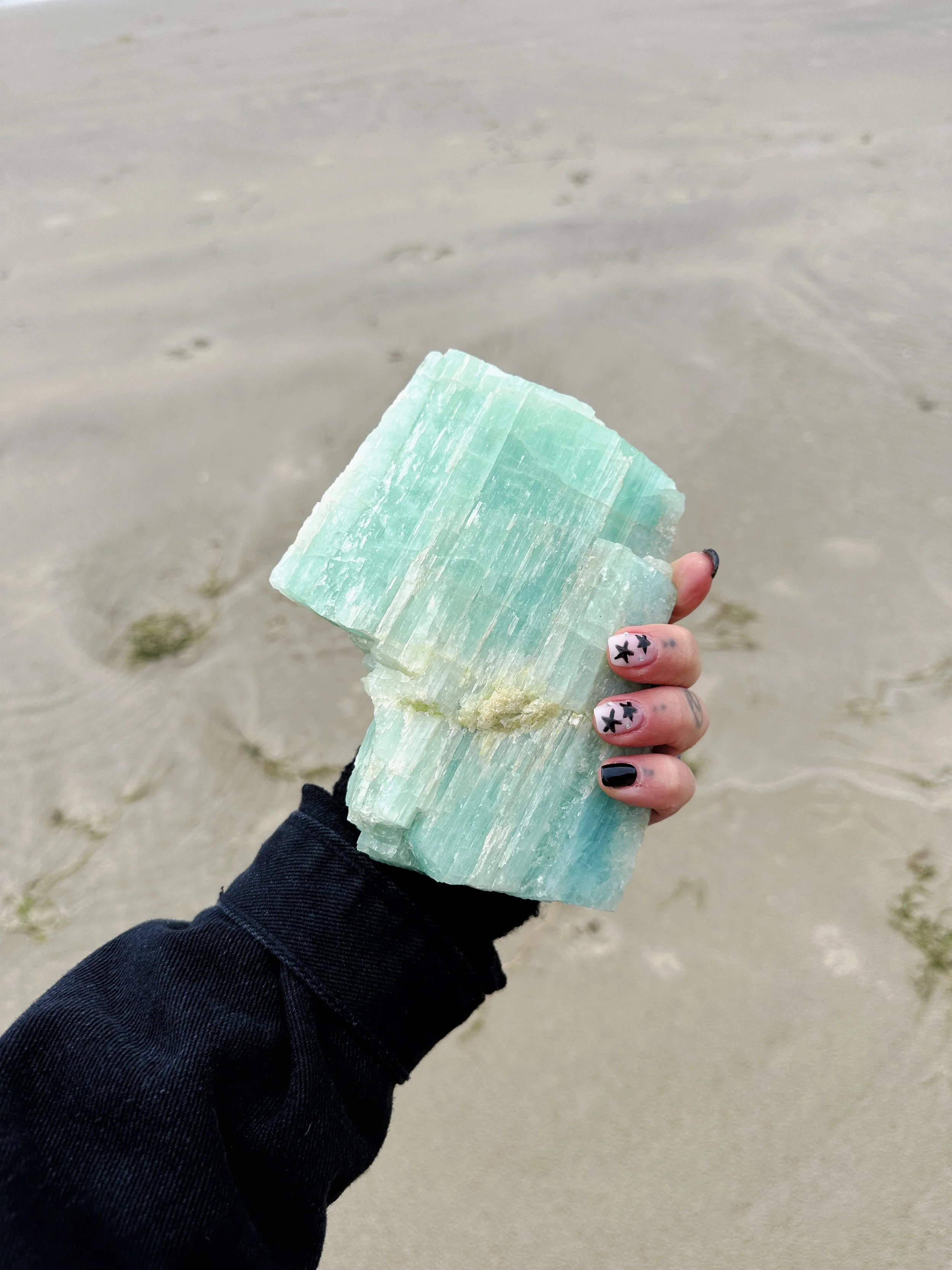 Person holding large green and white mineral or salt crystal on a beach with sand in the background.