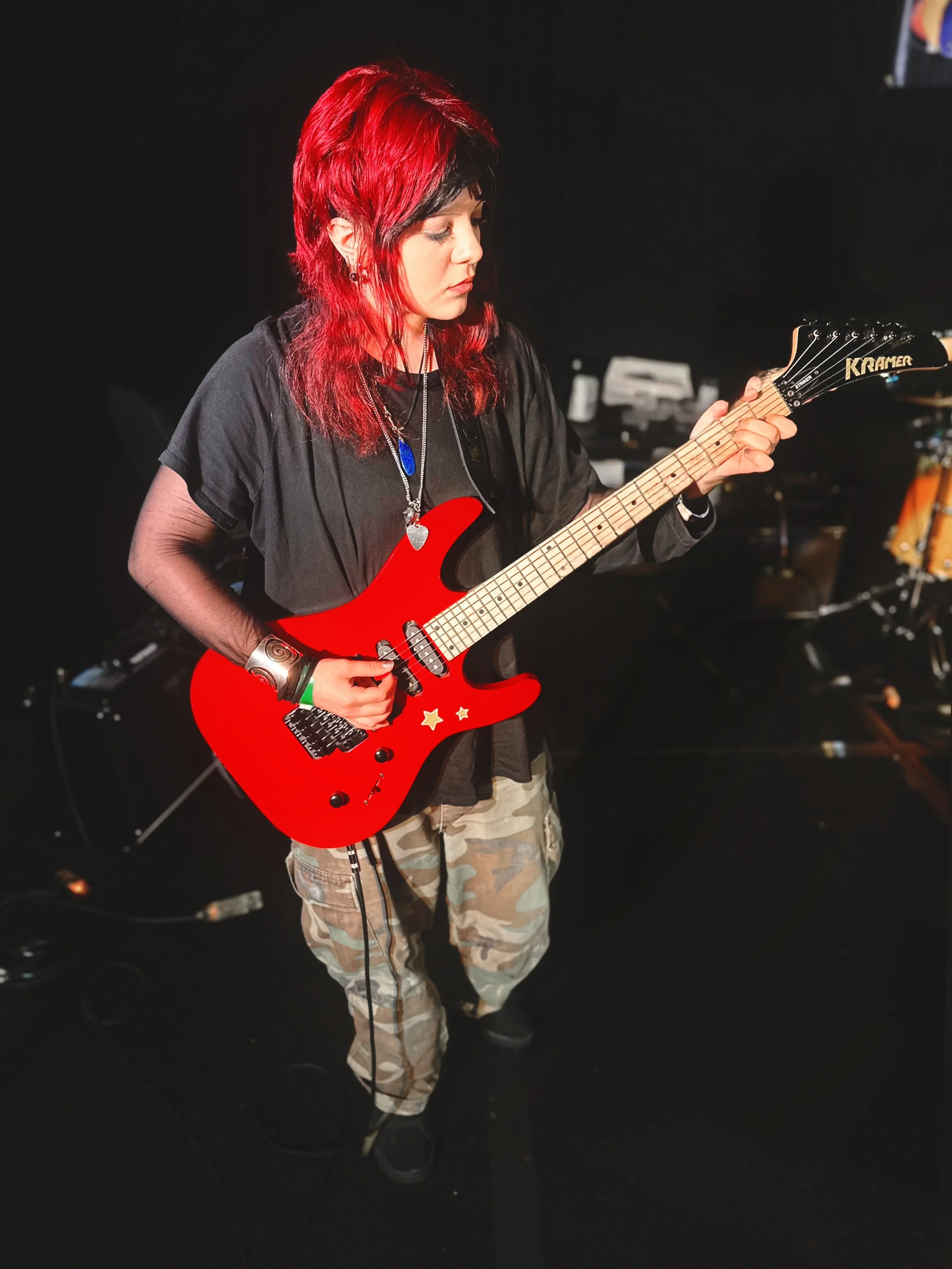 A woman with red and black hair playing a red electric guitar, wearing a black shirt, camouflage pants, and accessories, in a music studio or stage setting with musical equipment in the background.