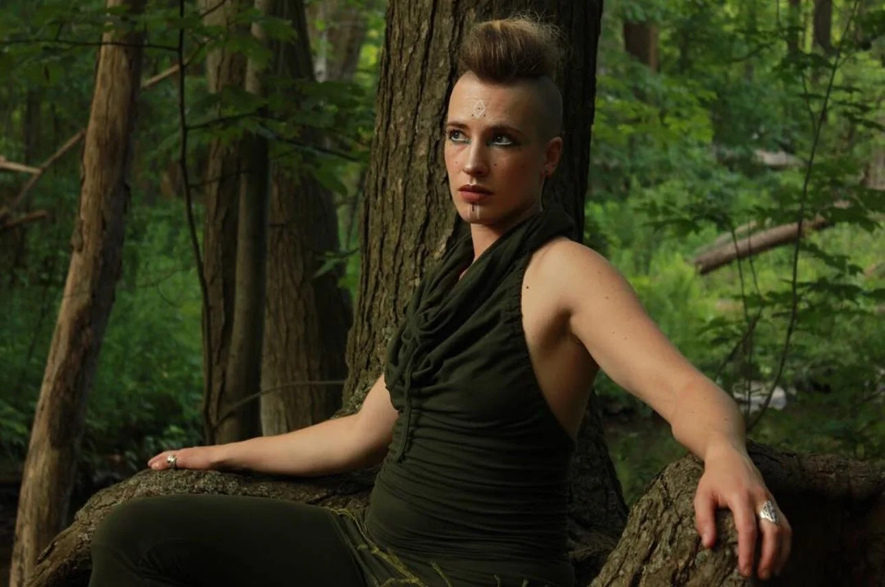 A woman with a mohawk hairstyle sitting against a tree in a wooded area, wearing a sleeveless dark green top and dark pants.
