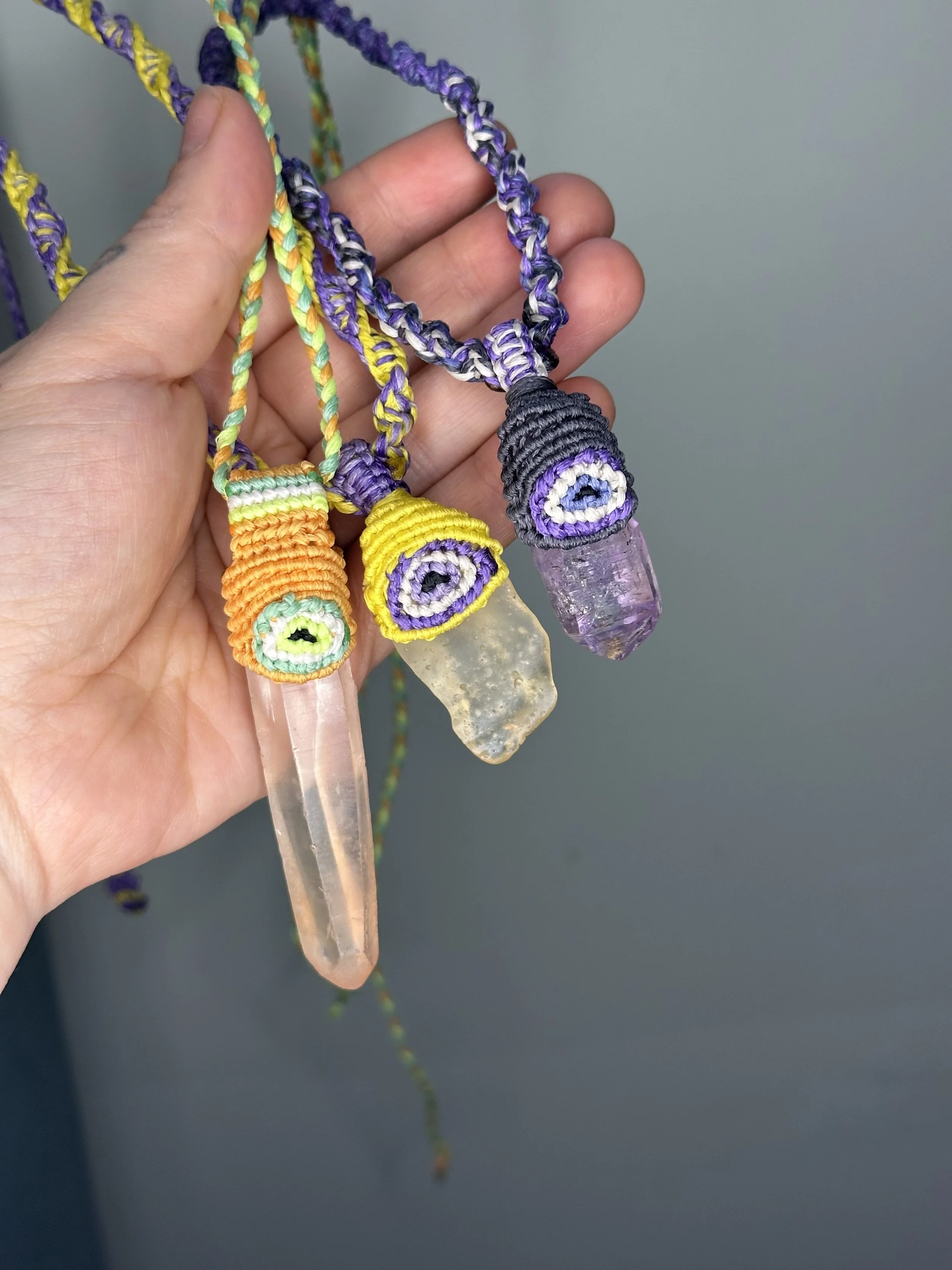 A person holding colorful handmade macrame necklaces with crystal pendants against a plain background.