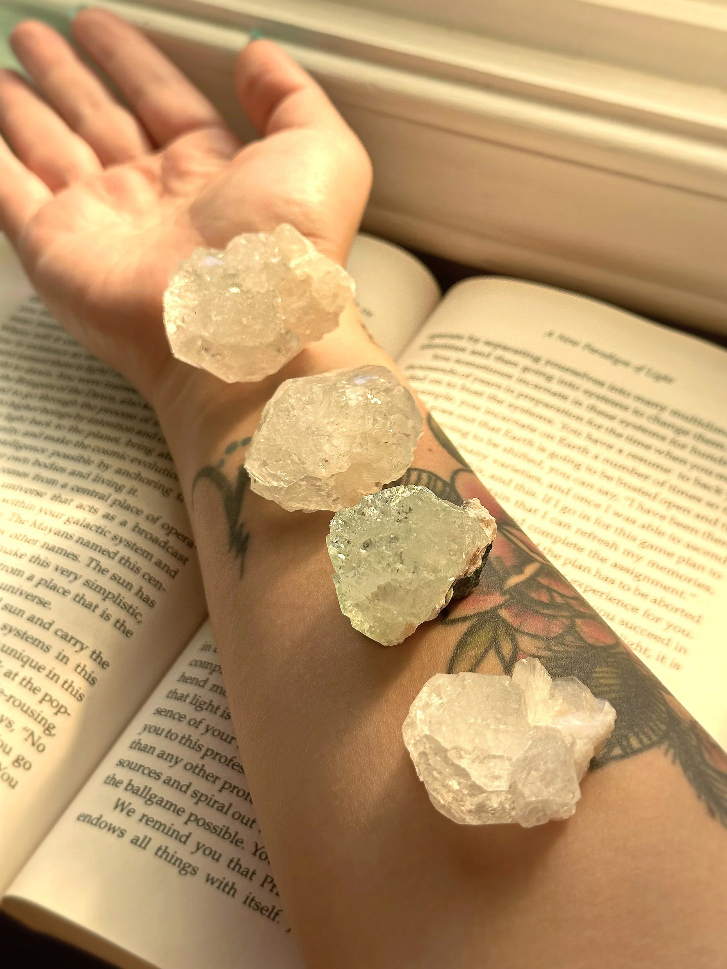 A person's arm with tattoos resting on an open book, with several large, clear crystals placed along the arm and on the book.