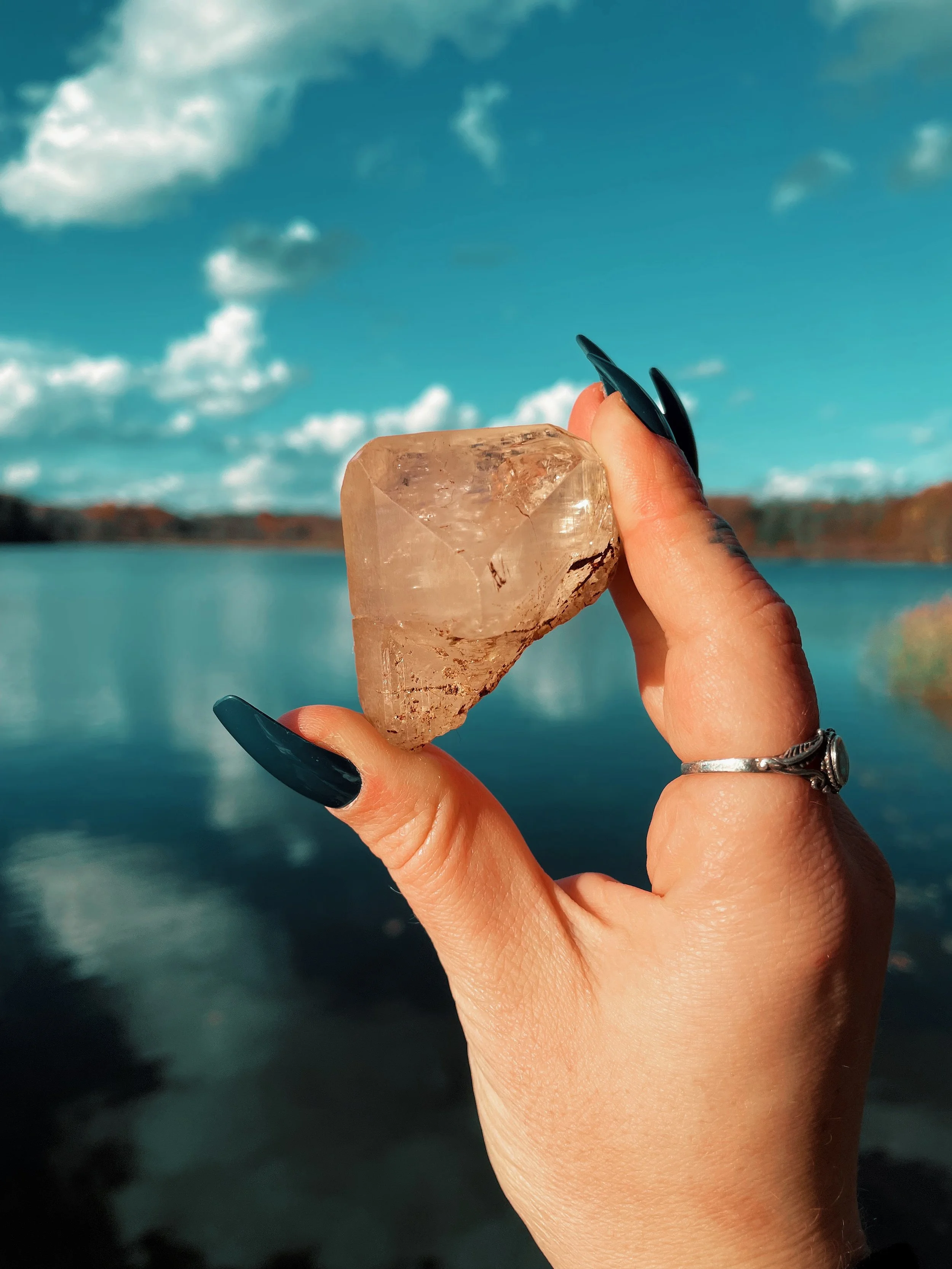 Person holding a large clear quartz crystal with a scenic lake and mountains in the background under a cloudy sky.