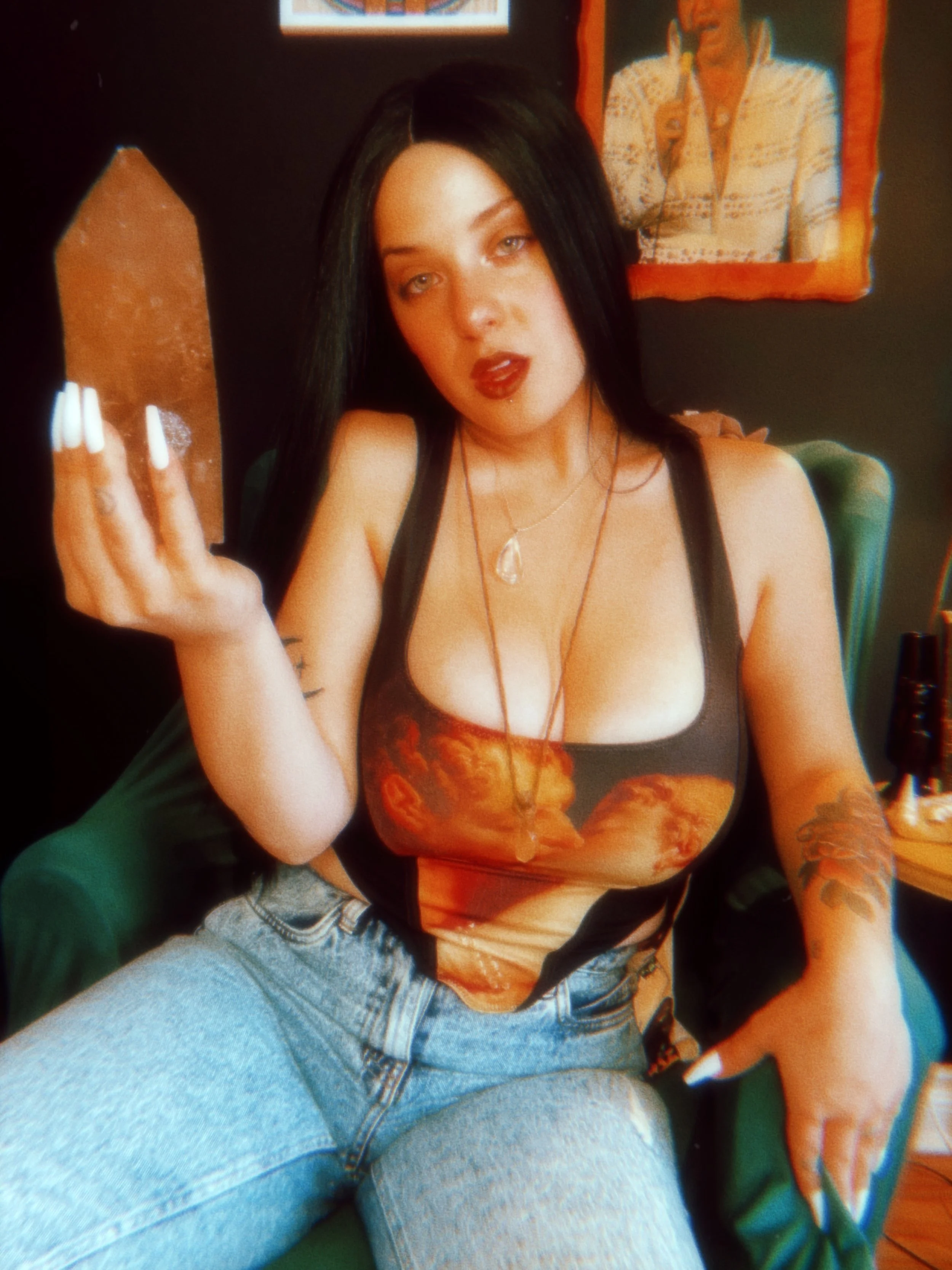 A woman with long black hair, wearing a tank top with a pizza print and light blue jeans, sitting on a chair. She has tattoos on her left arm, and is wearing necklaces and makeup with dark lipstick. She is holding a rectangular object in her right ha
