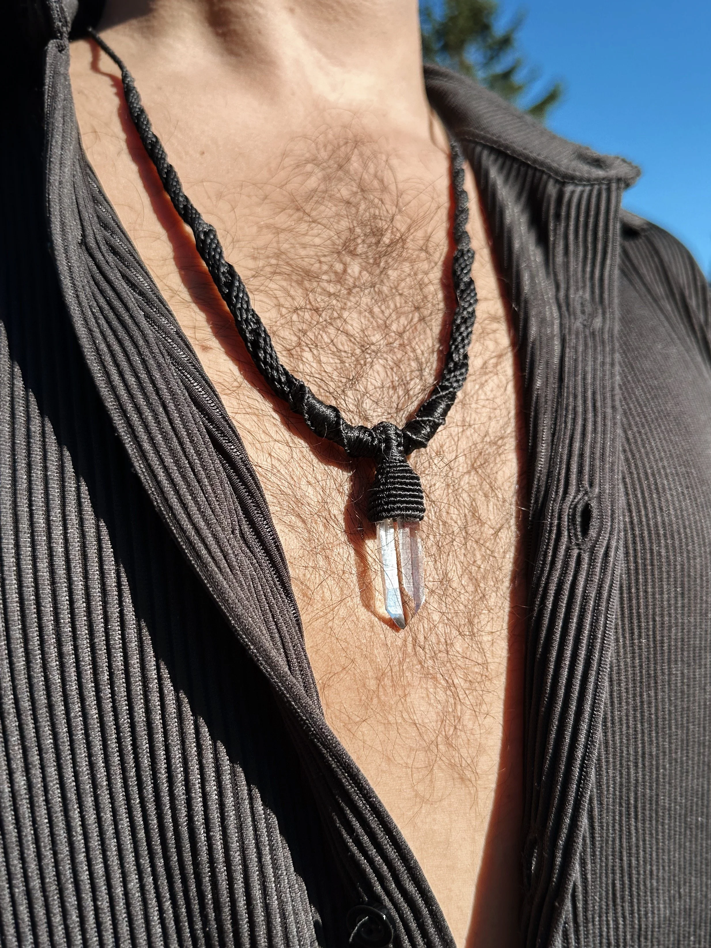 Close-up of a person's chest with chest hair, wearing a black cord necklace with a pointed crystal pendant and a black ribbed shirt unbuttoned.
