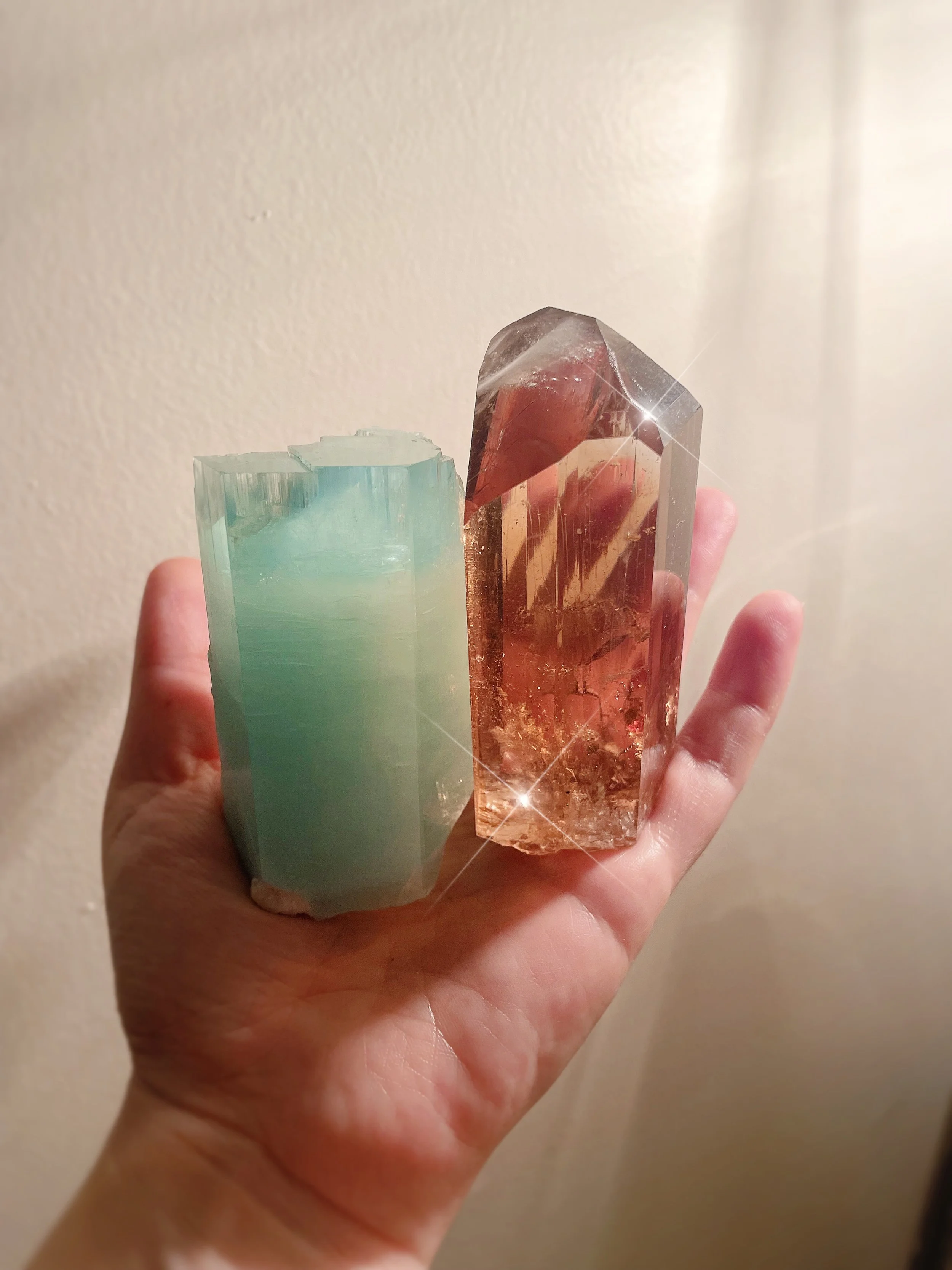 A hand holding three large, colorful crystals with a plain beige wall in the background.
