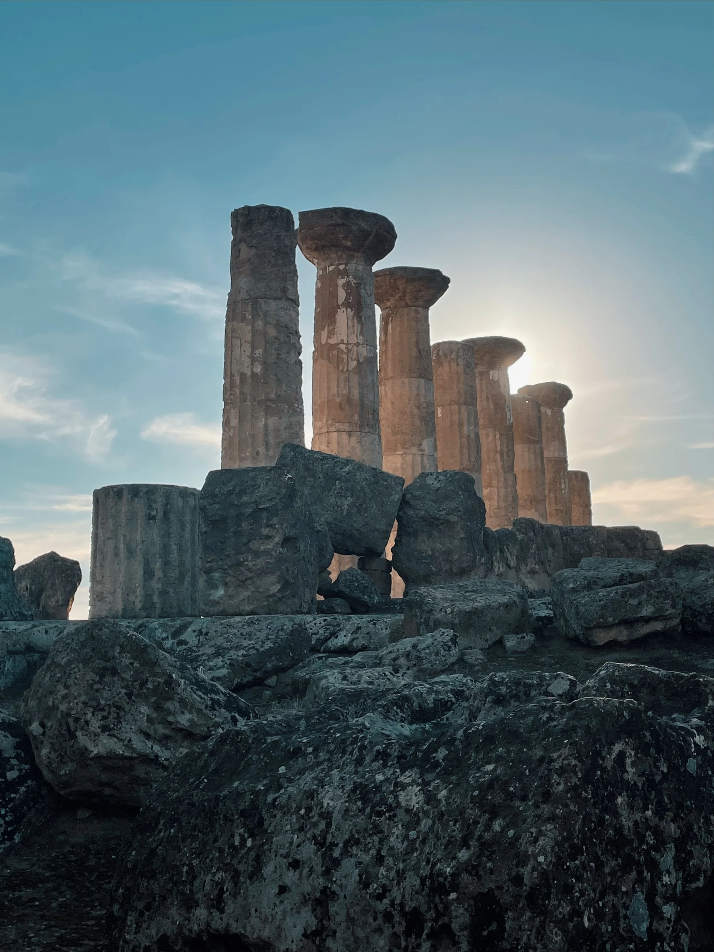 Ancient stone ruins with tall columns under a blue sky with the sun in the background.