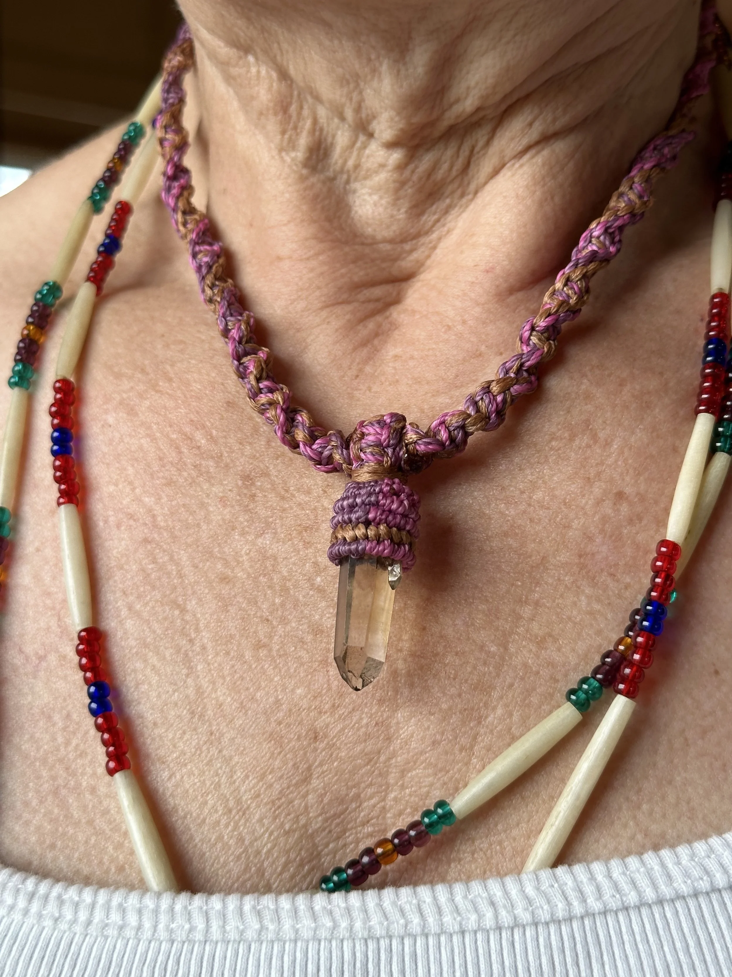 Close-up of a necklace with a clear quartz crystal pendant, wrapped in purple and pink thread, worn around a person's neck with multiple beaded necklaces.