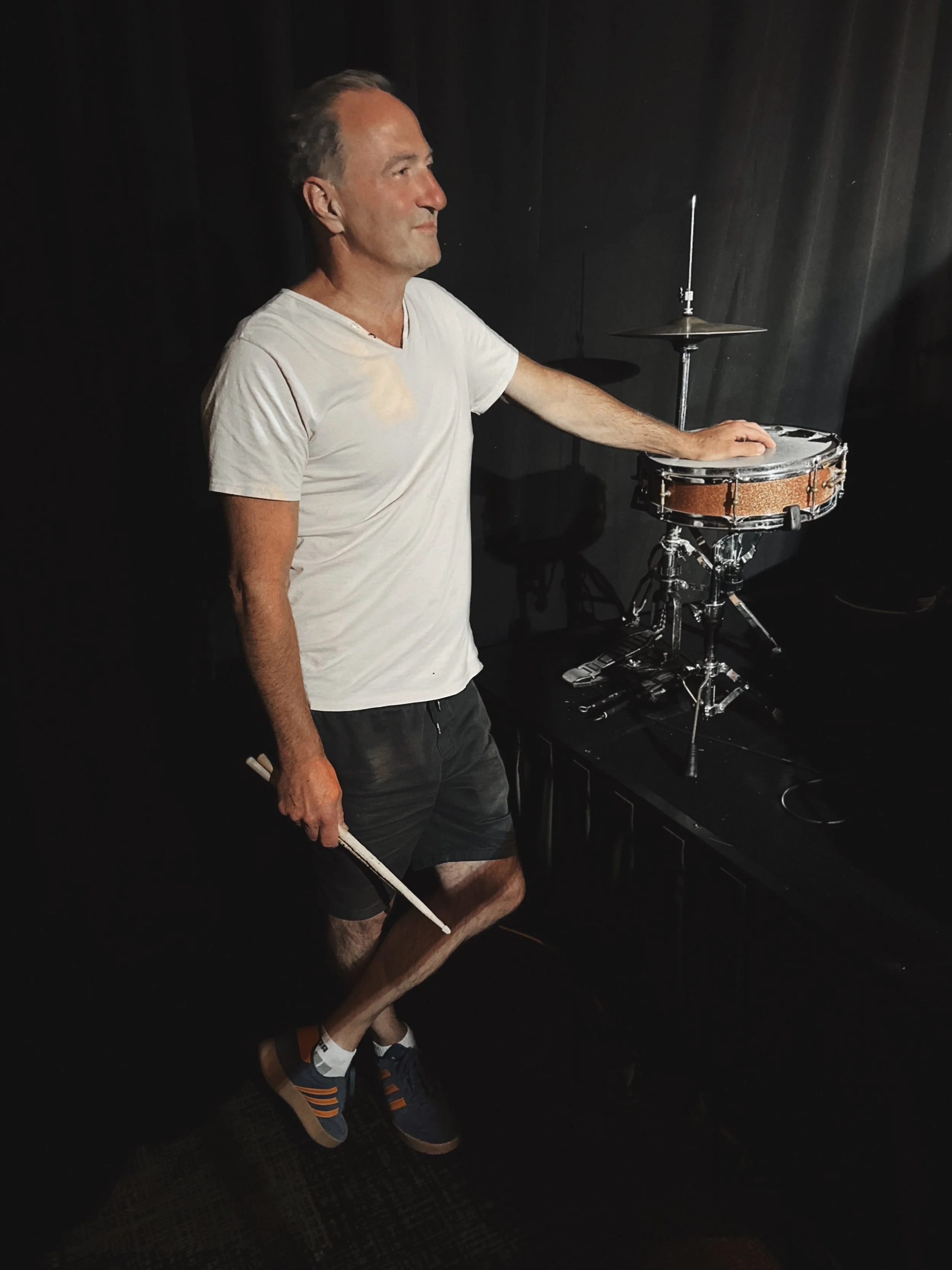 A man in a white t-shirt and black shorts stands next to a drum set in a dark room. He is holding a pair of drumsticks and has his hand resting on a small drum. The background is black and the floor has a dark checkered pattern.