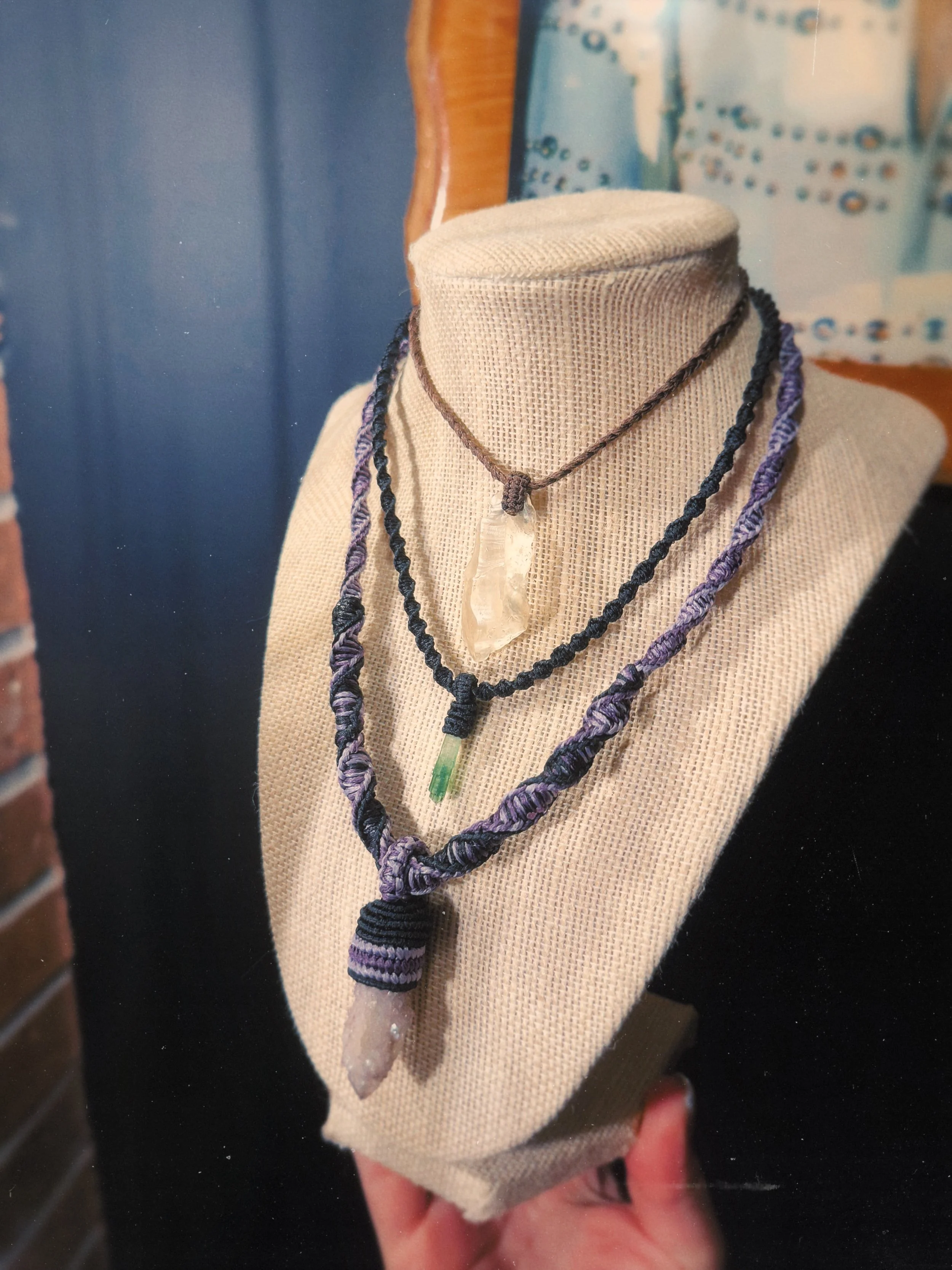 A person holding a small display bust with jewelry, including a clear quartz crystal pendant and two braided necklaces, one purple and black and another purple and blue.