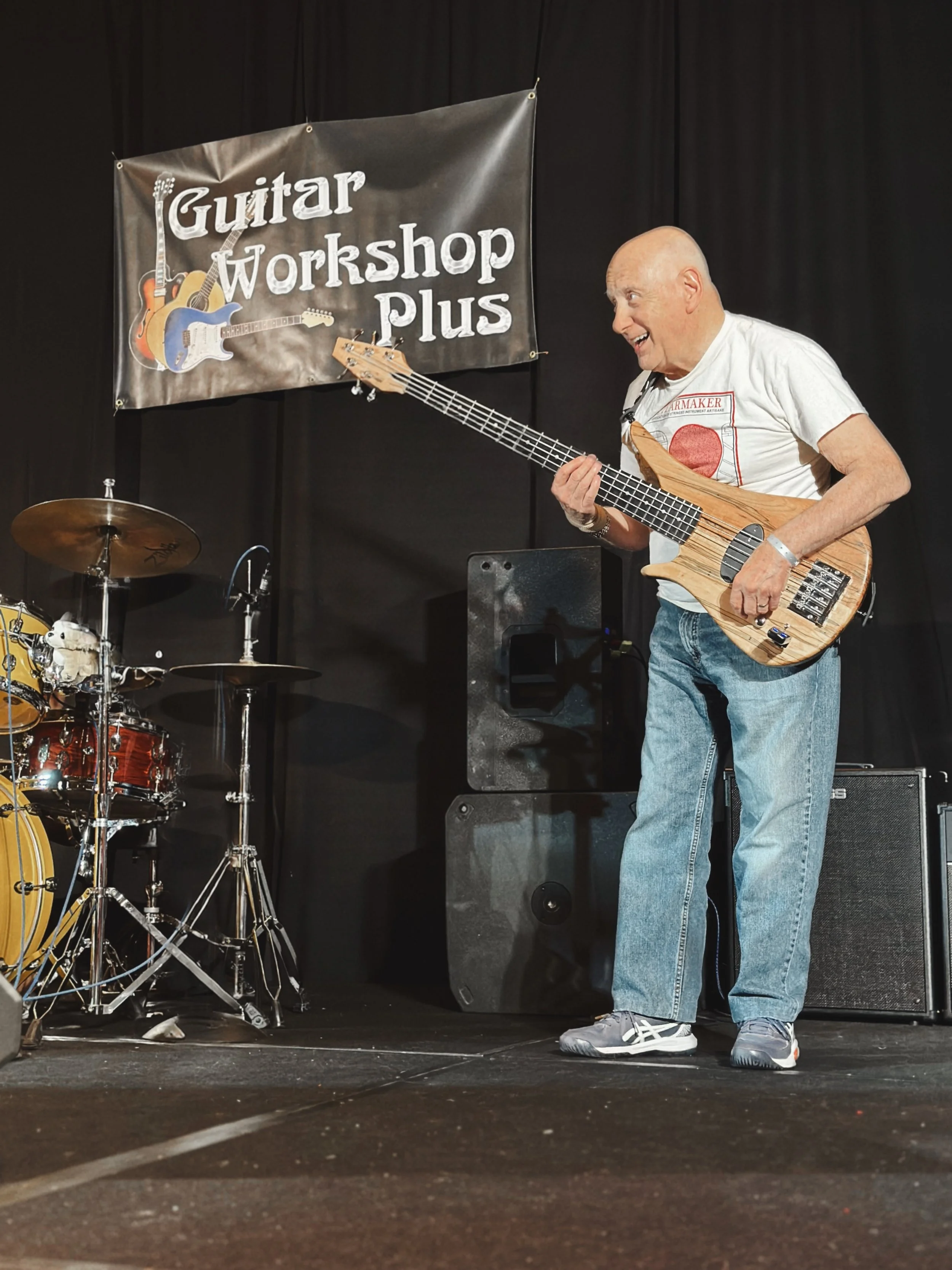 A man playing a bass guitar on stage at a guitar workshop event. There is a black backdrop with a sign that reads 'Guitar Workshop Plus' and pictures of guitars. The man appears to be enjoying himself, wearing casual clothing and sneakers.