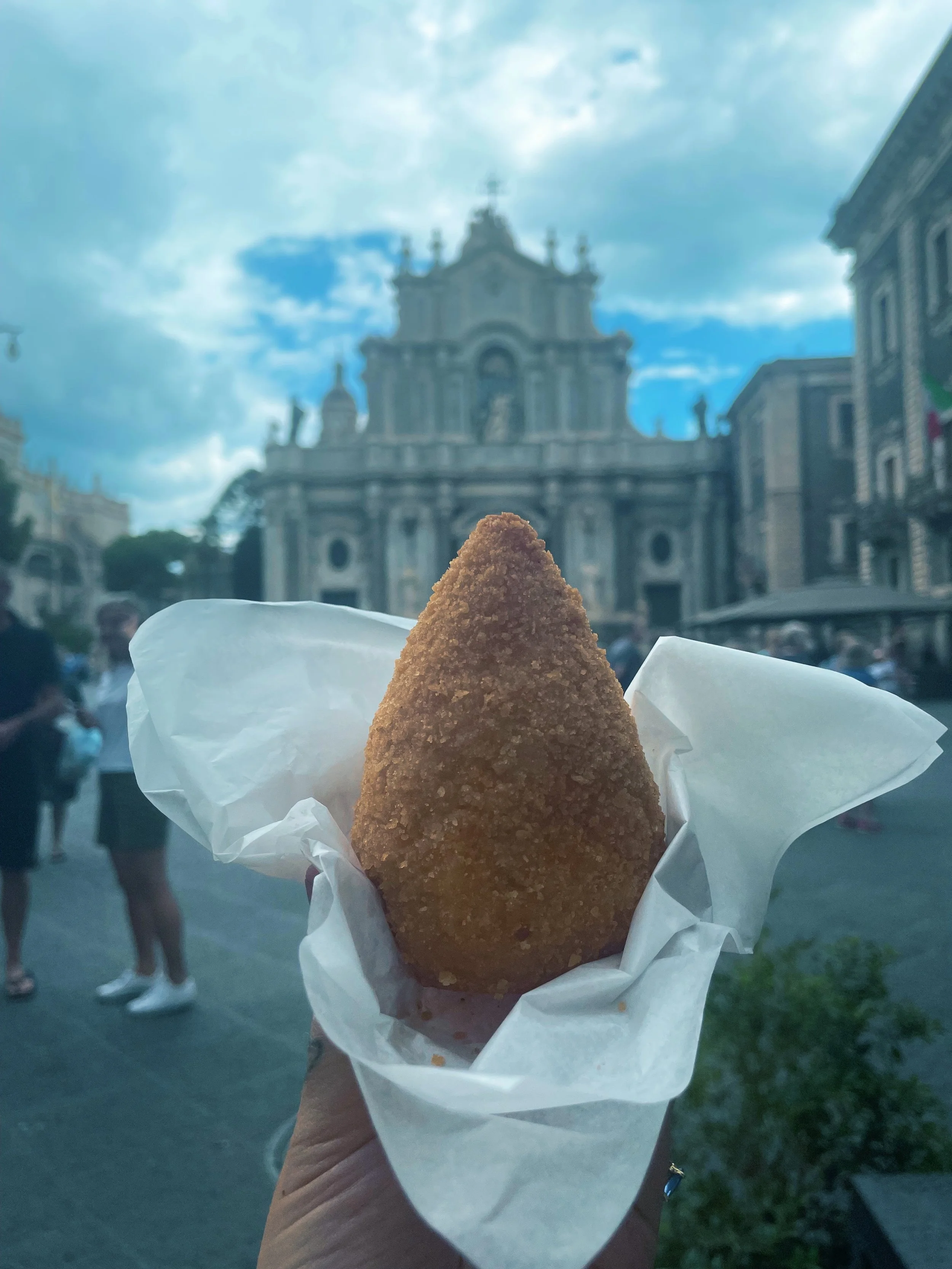 A person holding a fried food item wrapped in white paper in an outdoor setting, with a historic building with intricate architecture and another building visible in the background, under a partly cloudy sky.
