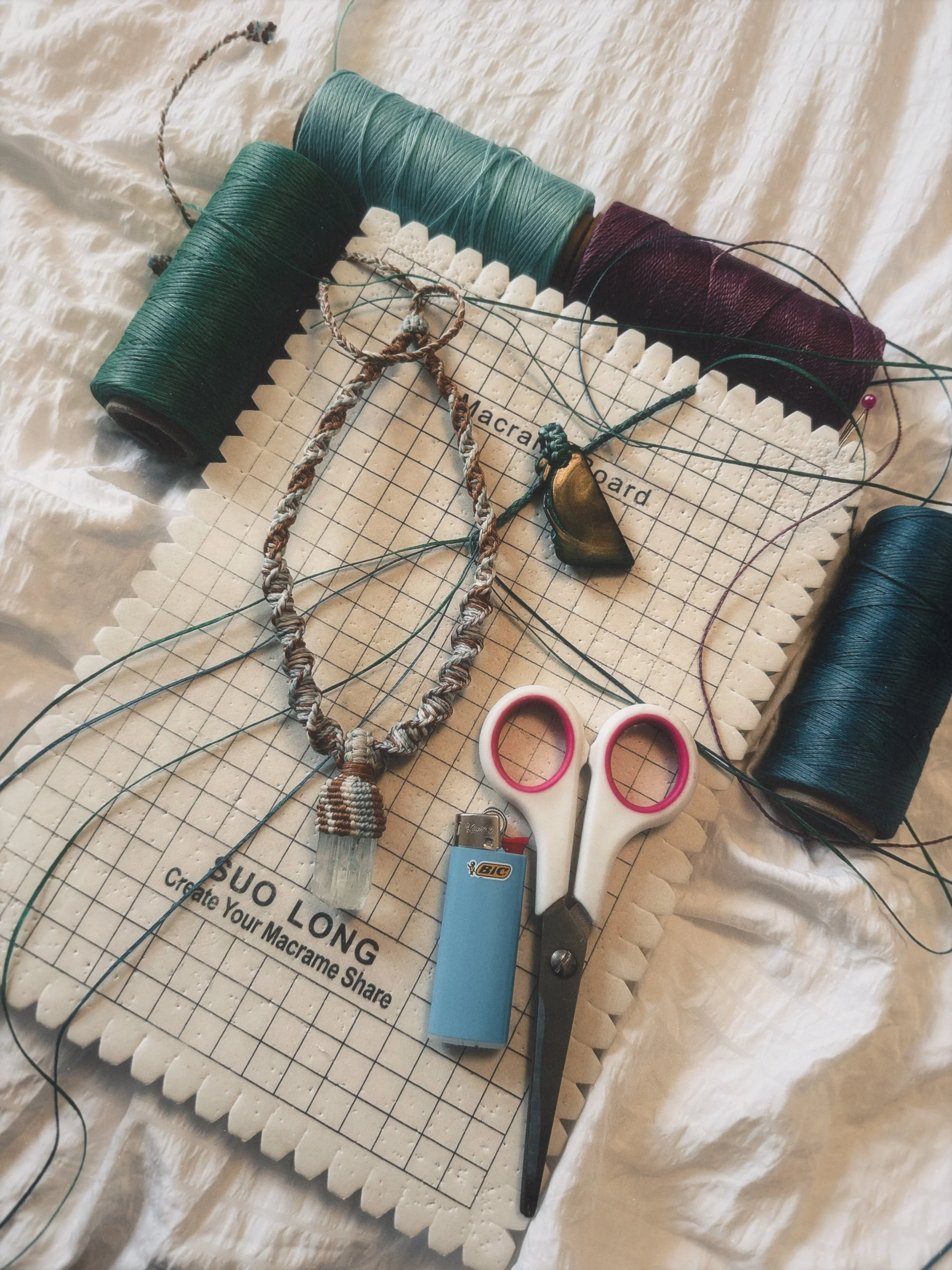 A workspace with rolls of green, teal, and burgundy thread, a macrame board, scissors, a lighter, a necklace with a gold-colored pendant, and various threading tools.