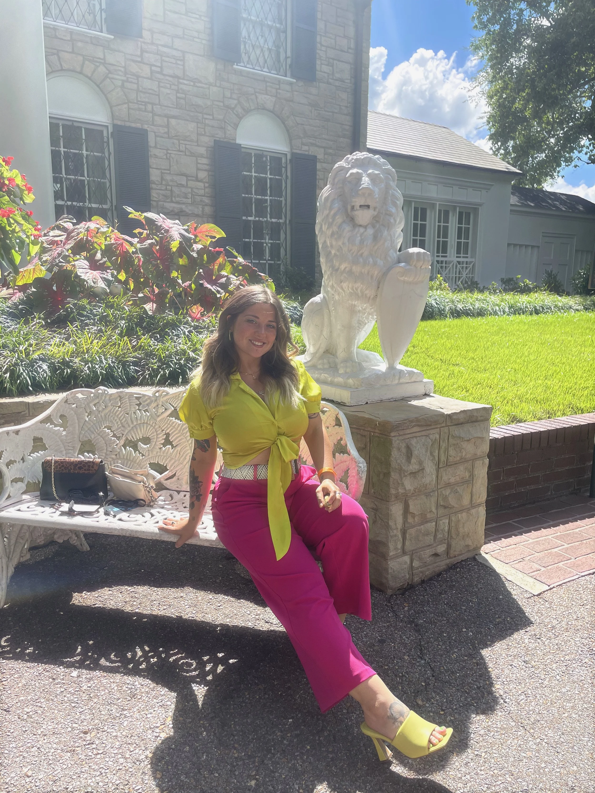 A woman sitting on a decorative white bench outdoors, wearing a bright yellow top and pink pants, with yellow high-heeled shoes, sitting near a white lion statue with a shield, in front of a brick house with blue shutters. She is smiling and has tattoos on her arm.