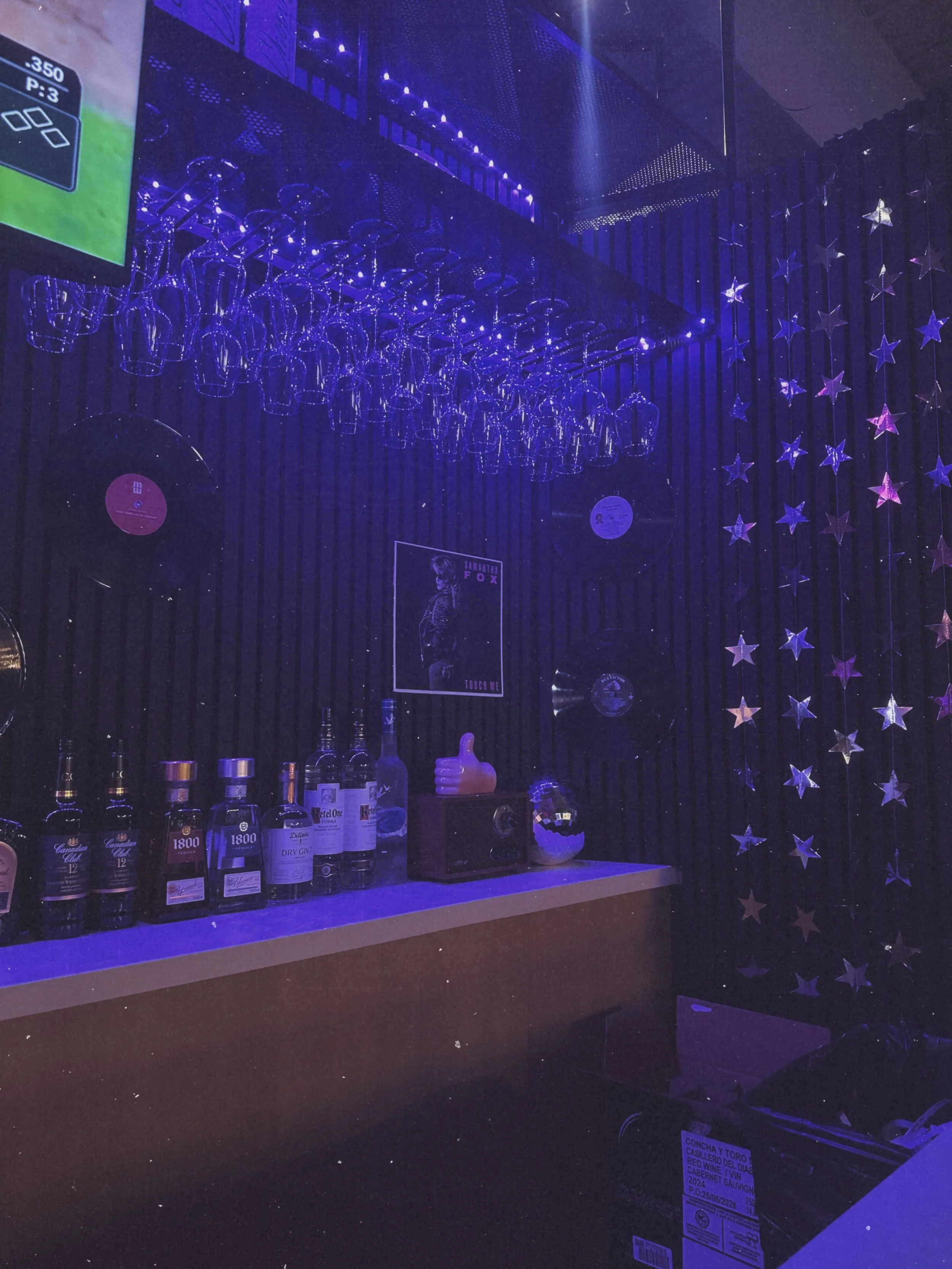 A bar with shelves of various bottles of alcohol, including whiskey, gin, and wine, illuminated by purple and blue lighting. Glassware is hanging upside down above the bar, and decorative star-shaped lights are on the wall.