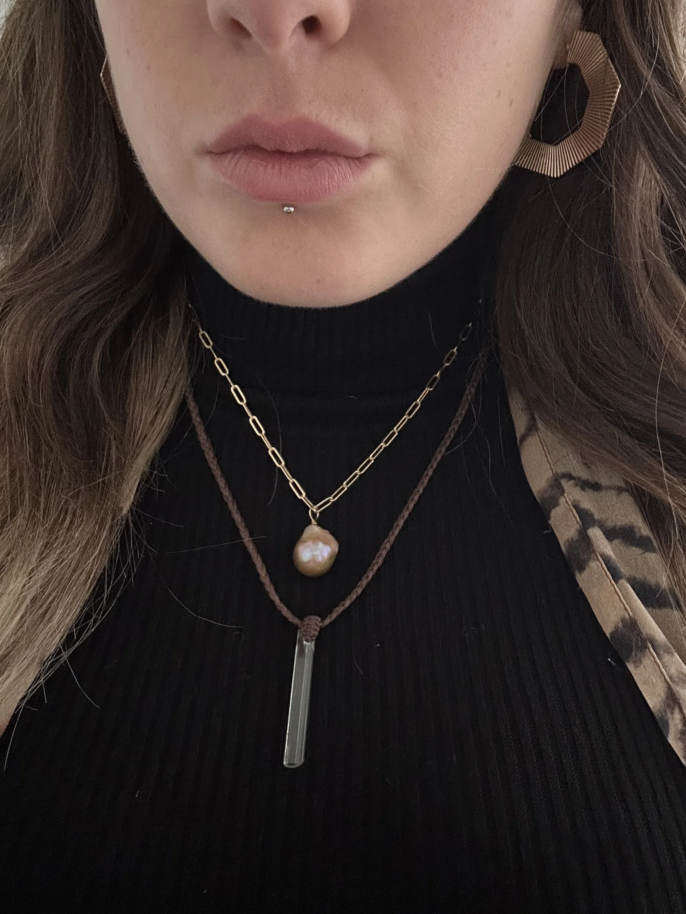 Close-up of a woman's lower face showing lips with a labret piercing, wearing layered necklaces with a pearl pendant, gold chain, and a silver bar pendant, and earrings with a black gemstone.