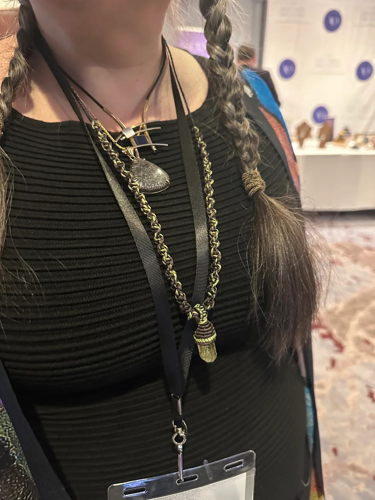 A person wearing three layered necklaces, featuring a dark stone pendant, a gold chain, and a handcrafted necklace with a yellow and brown pendant. They are dressed in a black ribbed top and have long hair braided on one side. There is a badge hangin