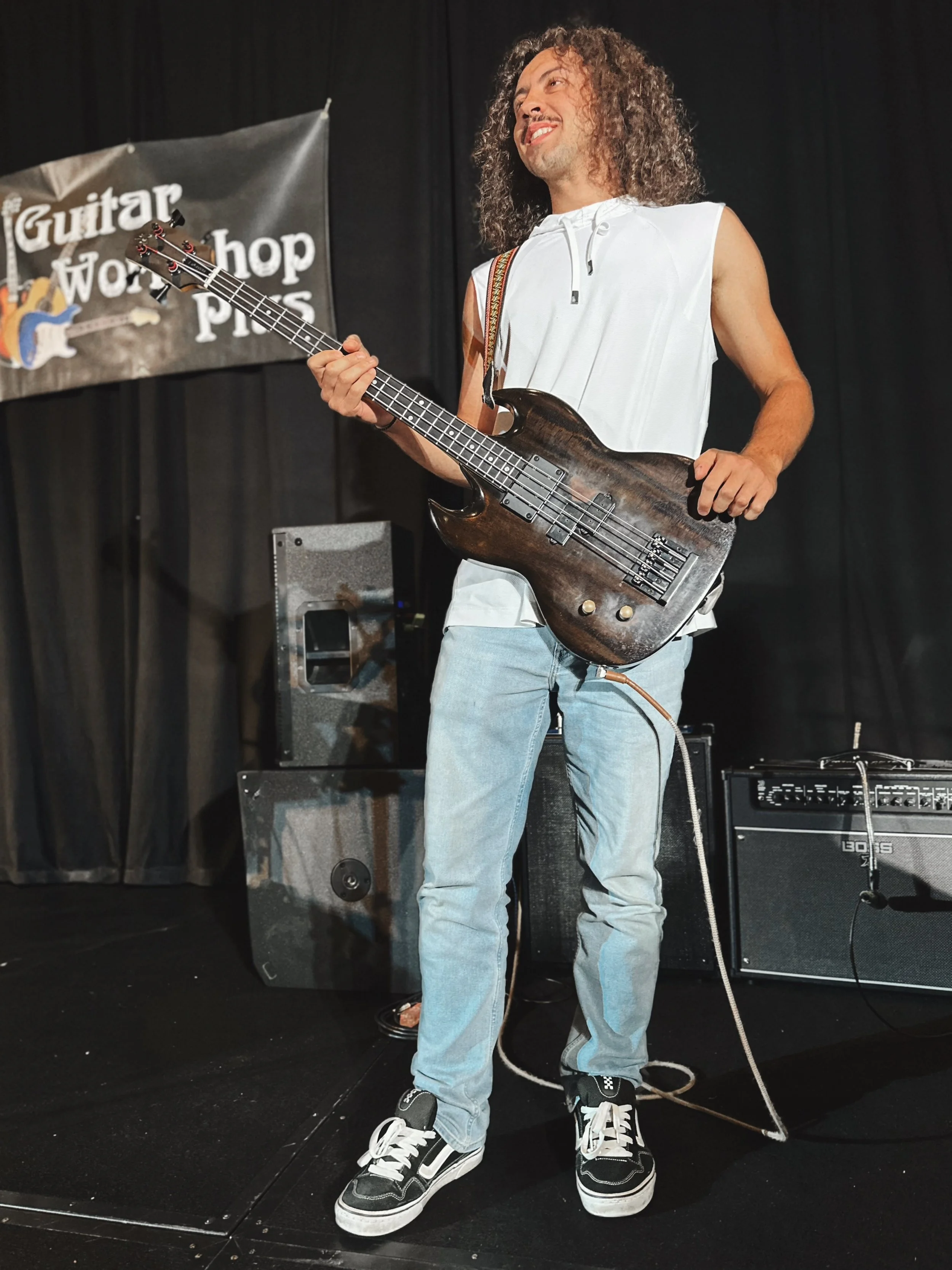 Young man with curly hair playing bass guitar on stage, wearing a sleeveless white hoodie, light blue jeans, and black and white sneakers, with music equipment in the background.