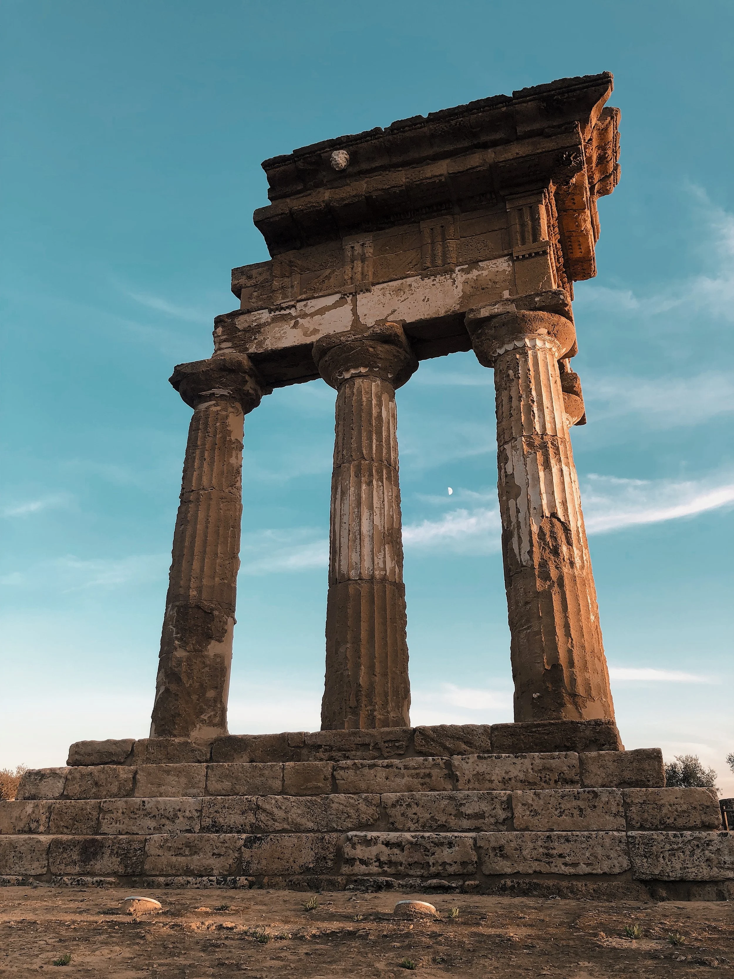 Ancient stone ruins with three tall, weathered columns supporting a horizontal stone structure, set against a blue sky with a visible moon in the background.