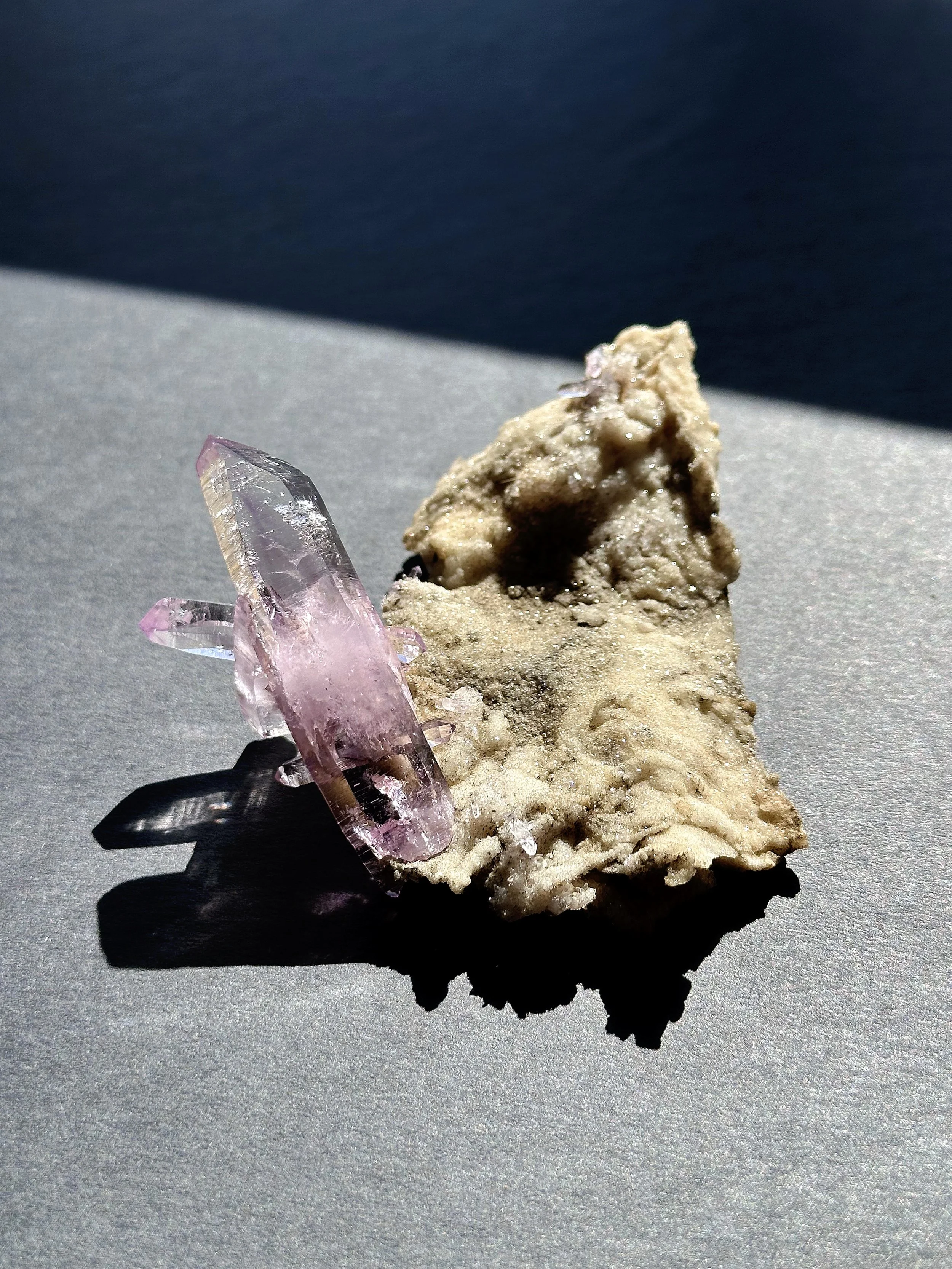 A pinkish-purple crystal with multiple points and a rough base, placed next to a beige mineral or rock with an irregular shape and textured surface, casting a shadow on a metallic surface.