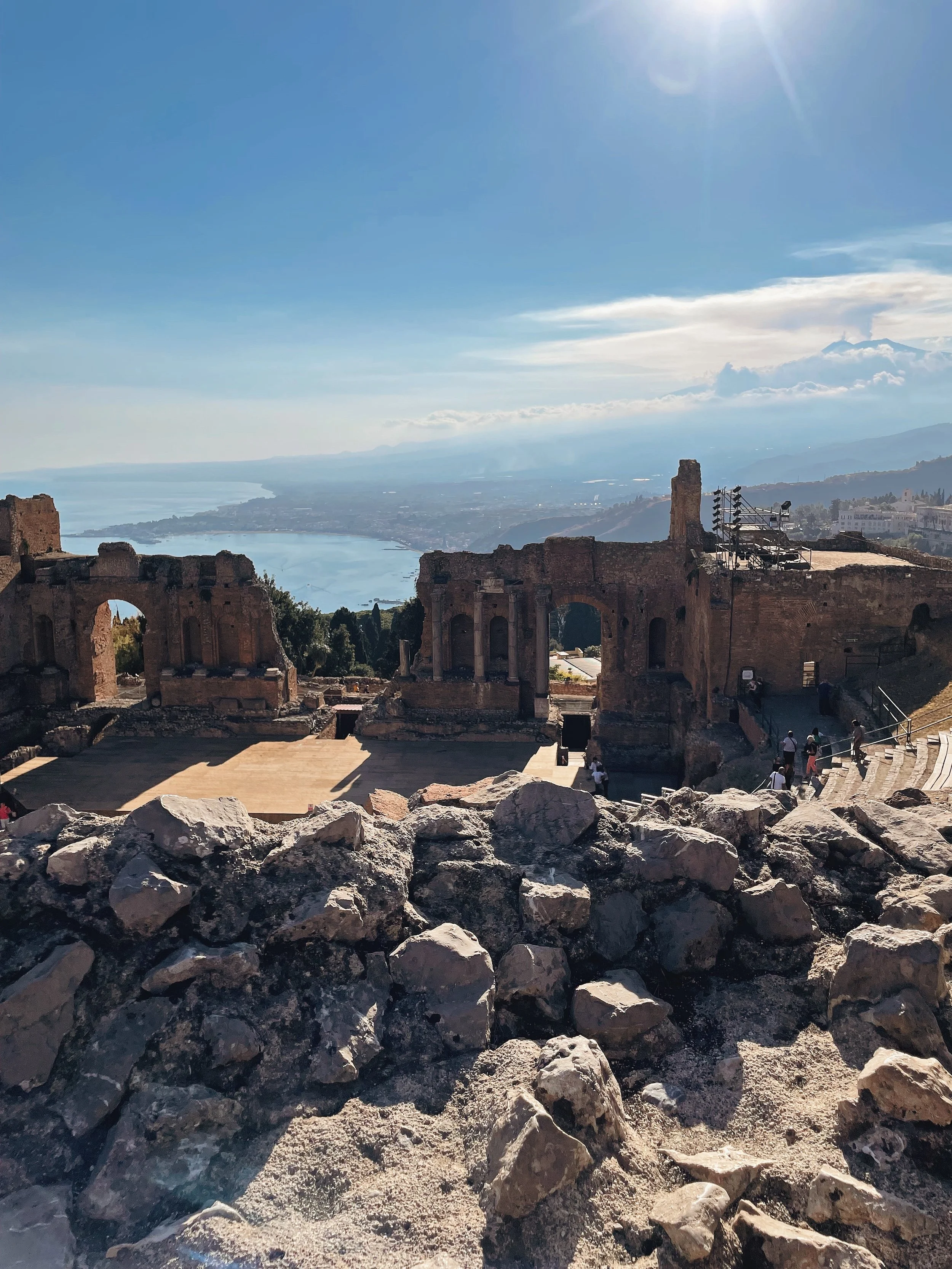 Ancient ruins with stone arches and walls overlooking a river and distant mountains under a clear blue sky with bright sunlight.