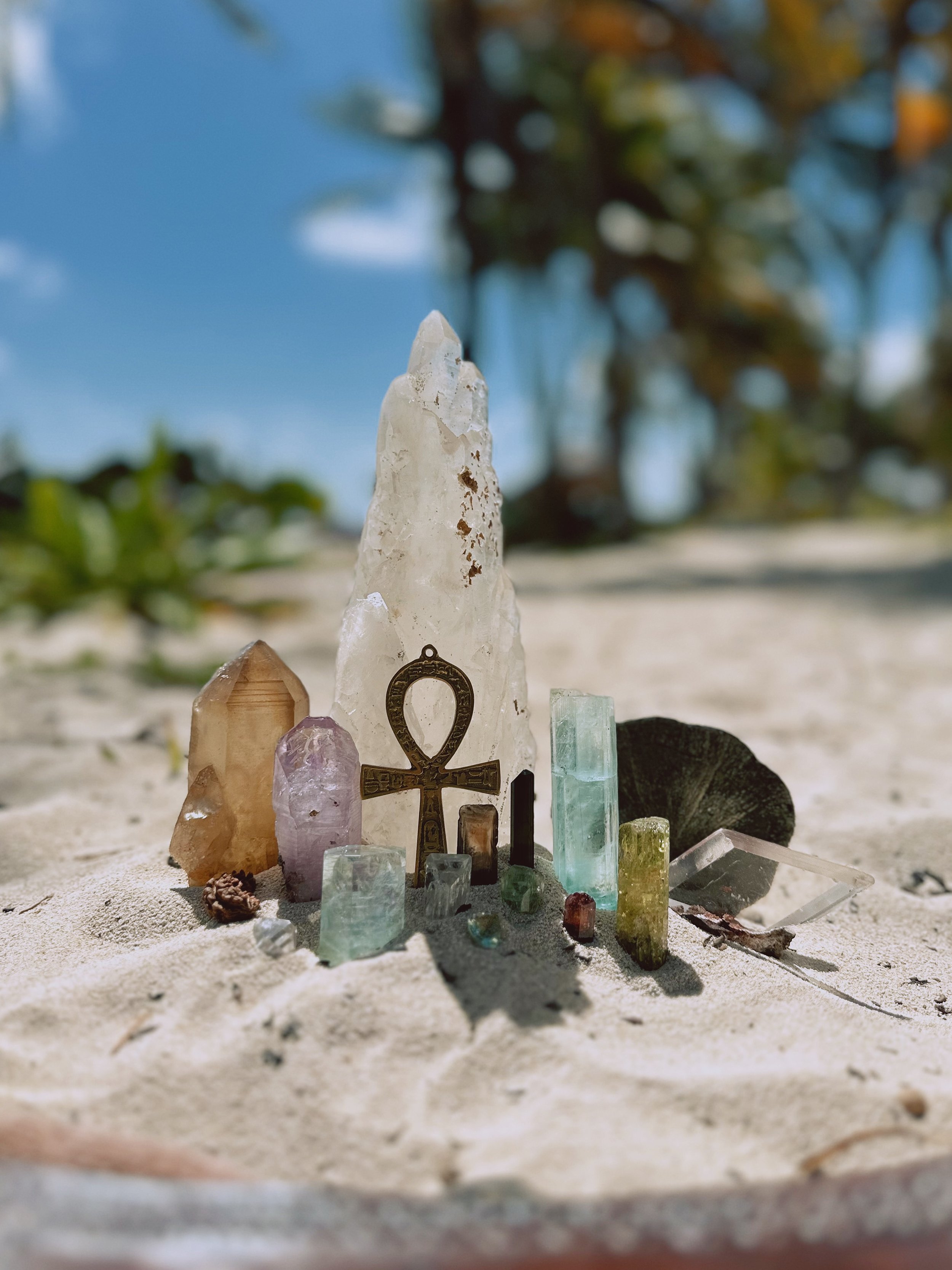 Collection of crystals and minerals placed on sandy ground outdoors, with blurred trees and blue sky in the background.