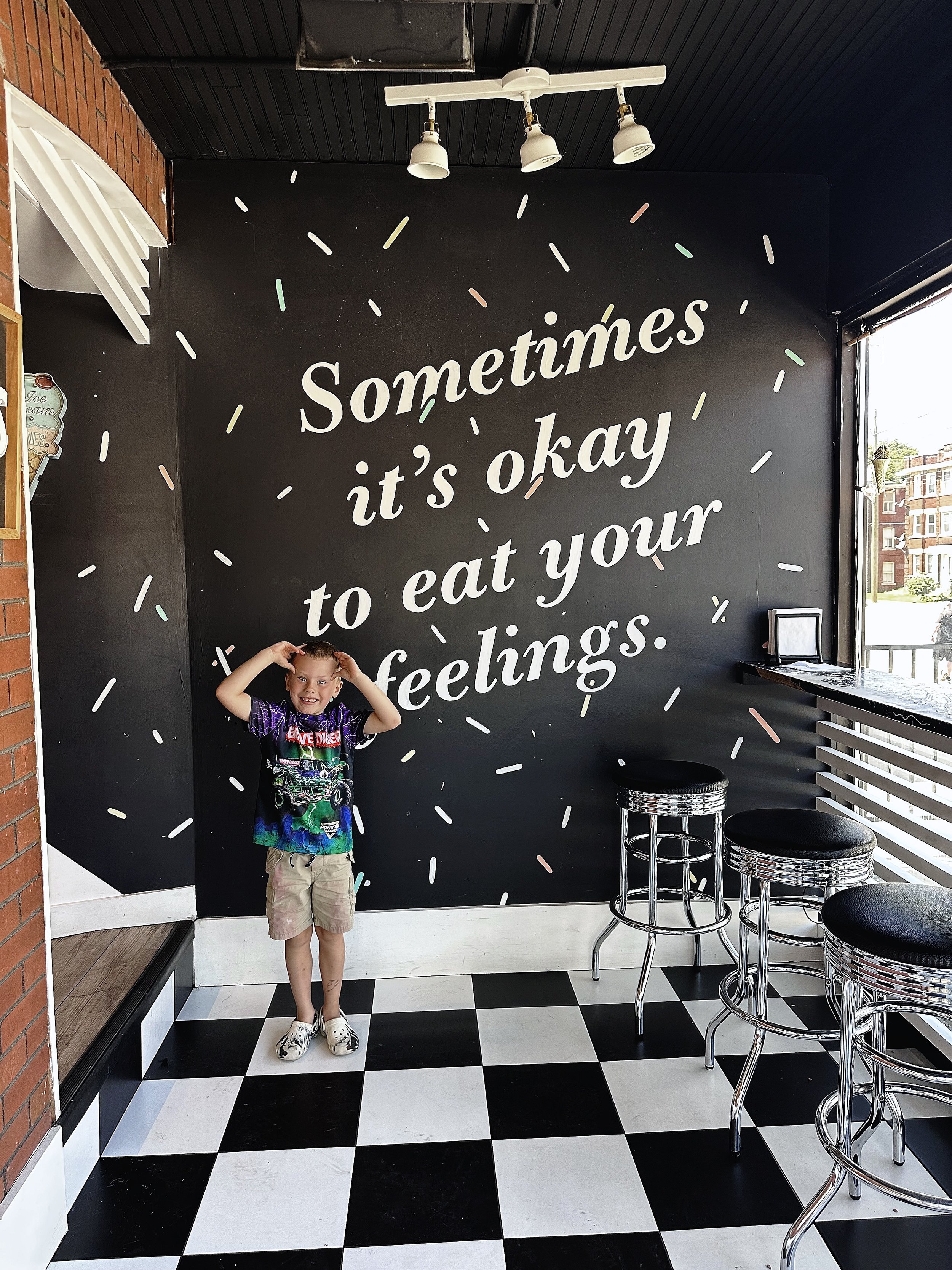 A young boy standing inside a black and white checkered floor restaurant or cafe, smiling and touching his hair, with a black wall behind him that has white text saying, 'Sometimes it's okay to eat your feelings,' and some colorful sprinkles design a