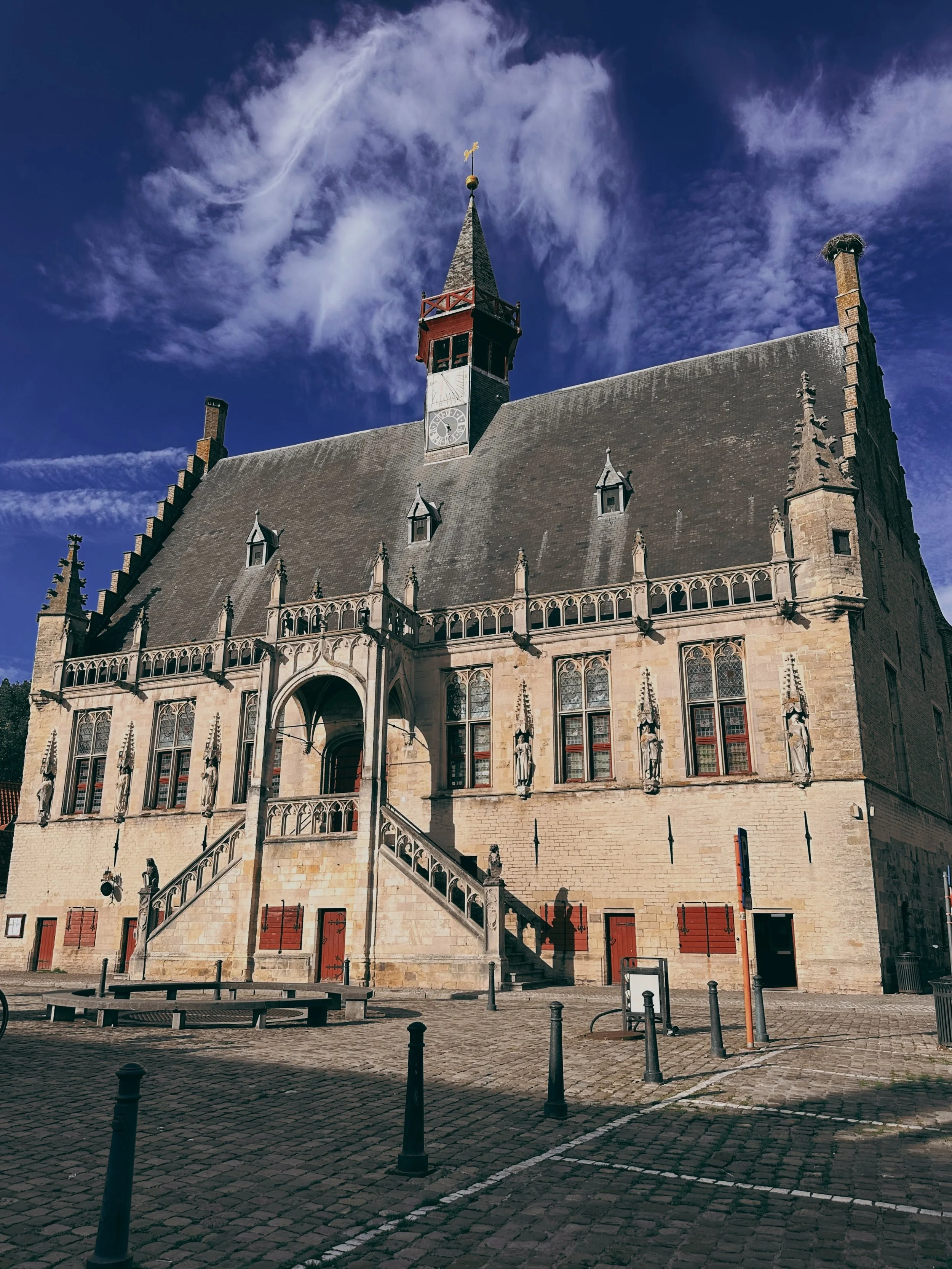 A historic stone building with a pointed roof, clock, and statues in front, situated in a cobblestone square with black posts and orange safety barriers, under a bright, partly cloudy sky.