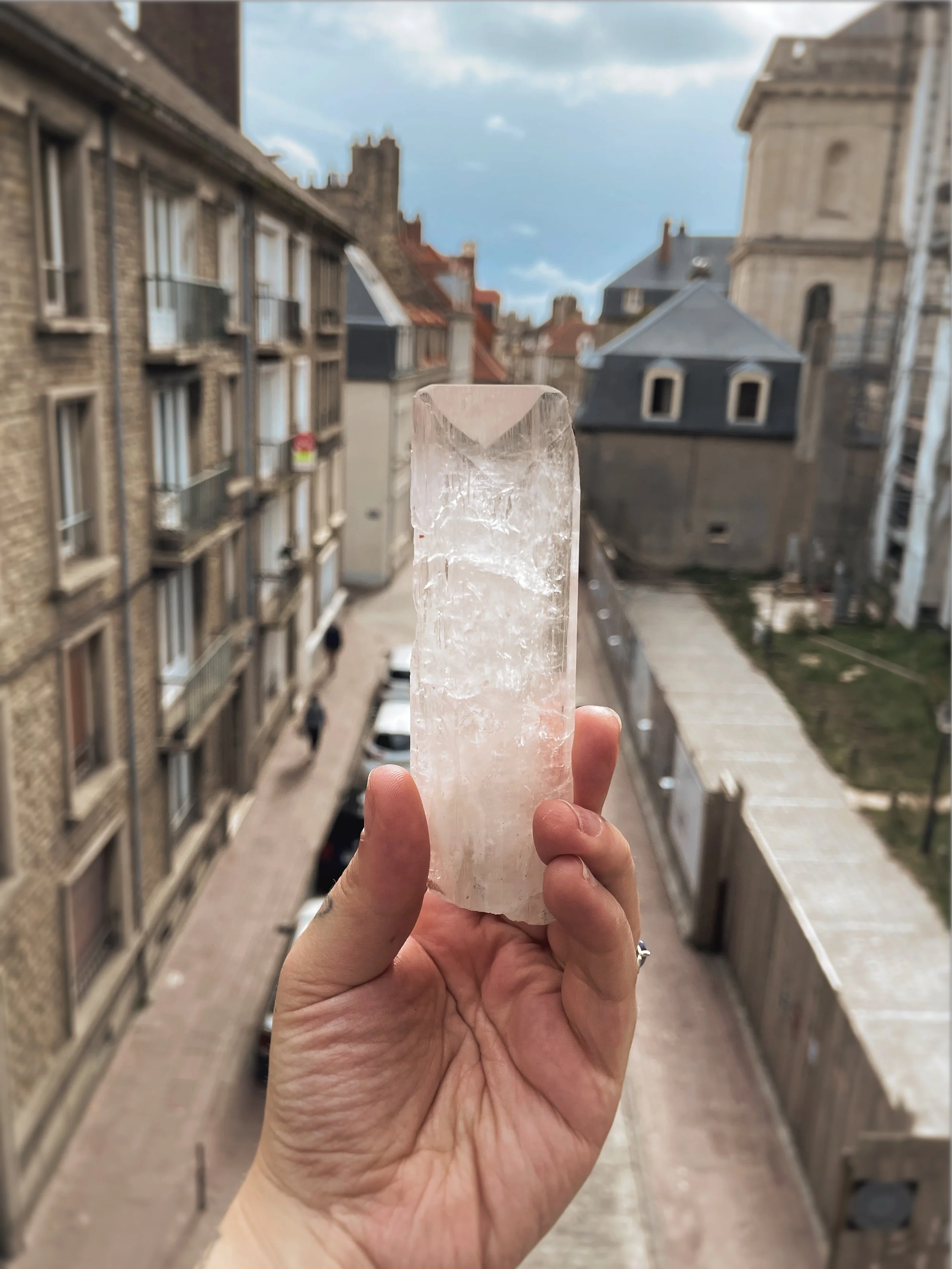 A hand holding a large, clear quartz crystal with city buildings and a partly cloudy sky in the background.