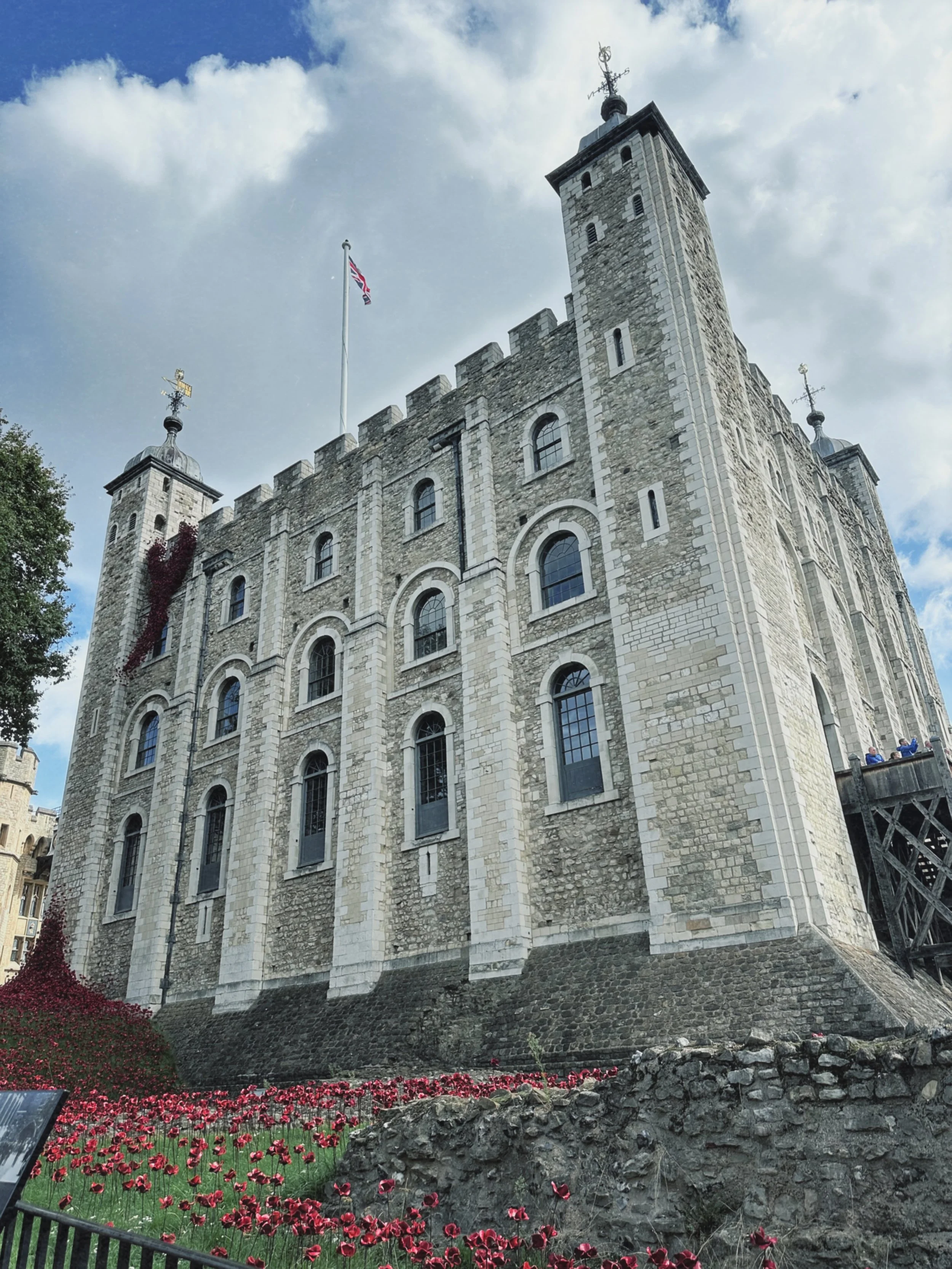 View of a tall historic stone castle with arched windows and multiple towers, topped with weather vanes and a Union Jack flag, against a partly cloudy sky. There are red flowers and greenery in the foreground.
