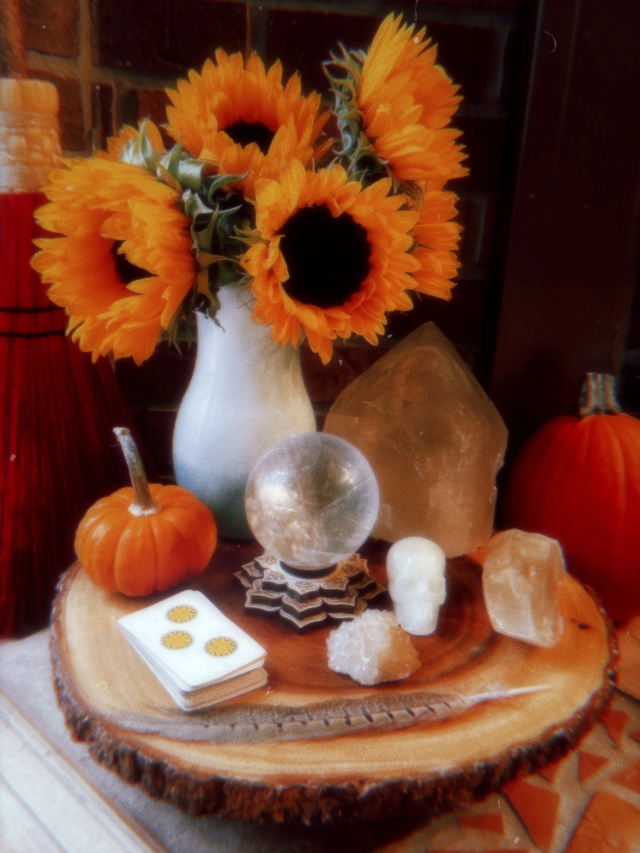 A tabletop display featuring a white vase with artificial orange sunflowers, small pumpkins, salt lamps, crystals, and a tarot card deck on a wooden slice.