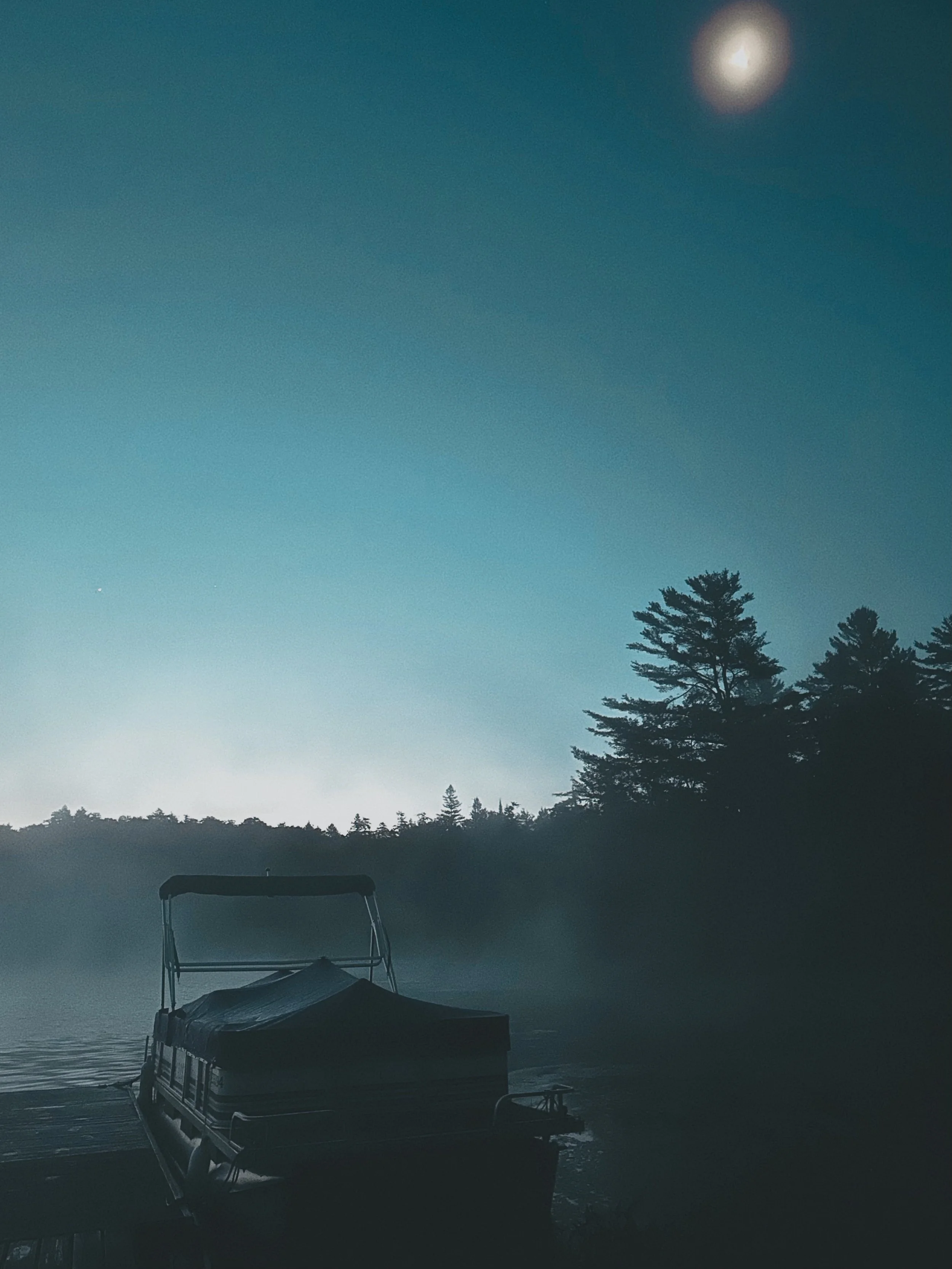 A boat docked at a wooden pier on a calm lake during dusk or dawn, with trees and hills in the background under a clear sky with the moon visible.