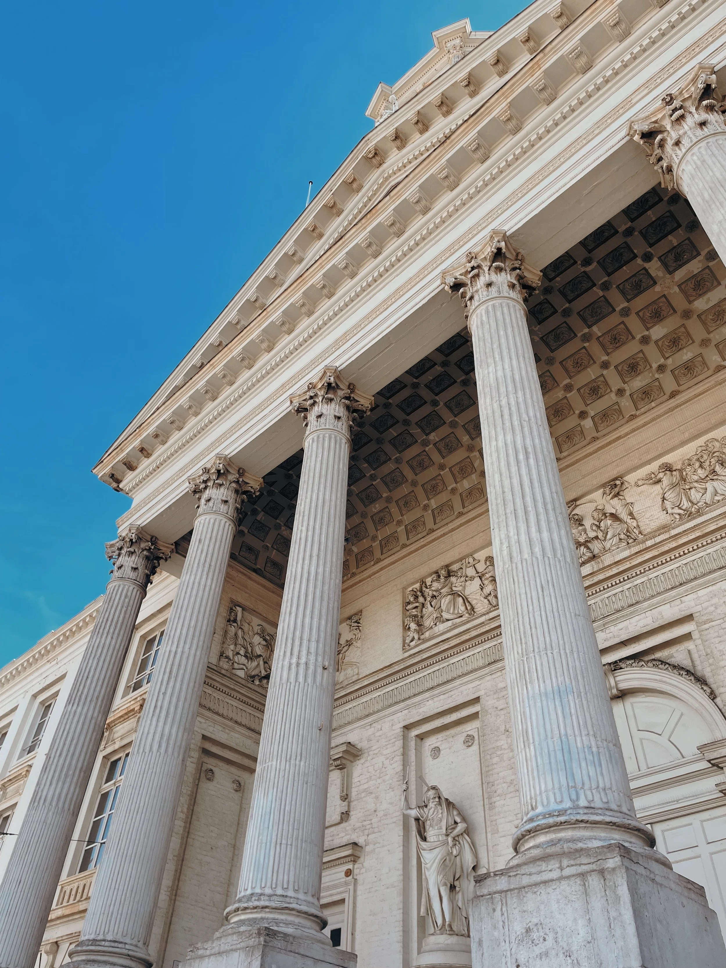 Close-up of classical Greek-style building with large stone columns and detailed sculptures on the facade under a clear blue sky.