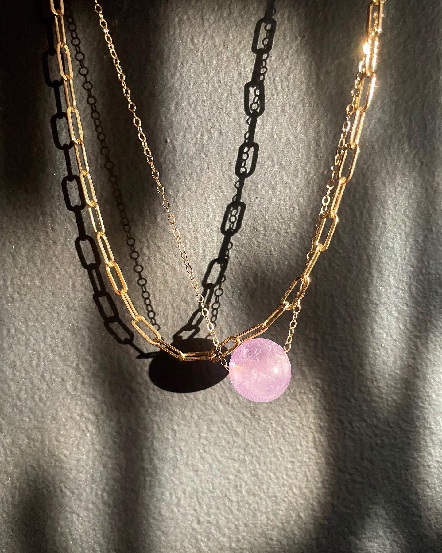 Three layered necklaces with gold and black chains, adorned with a pink circular pendant, displayed on a textured dark surface.