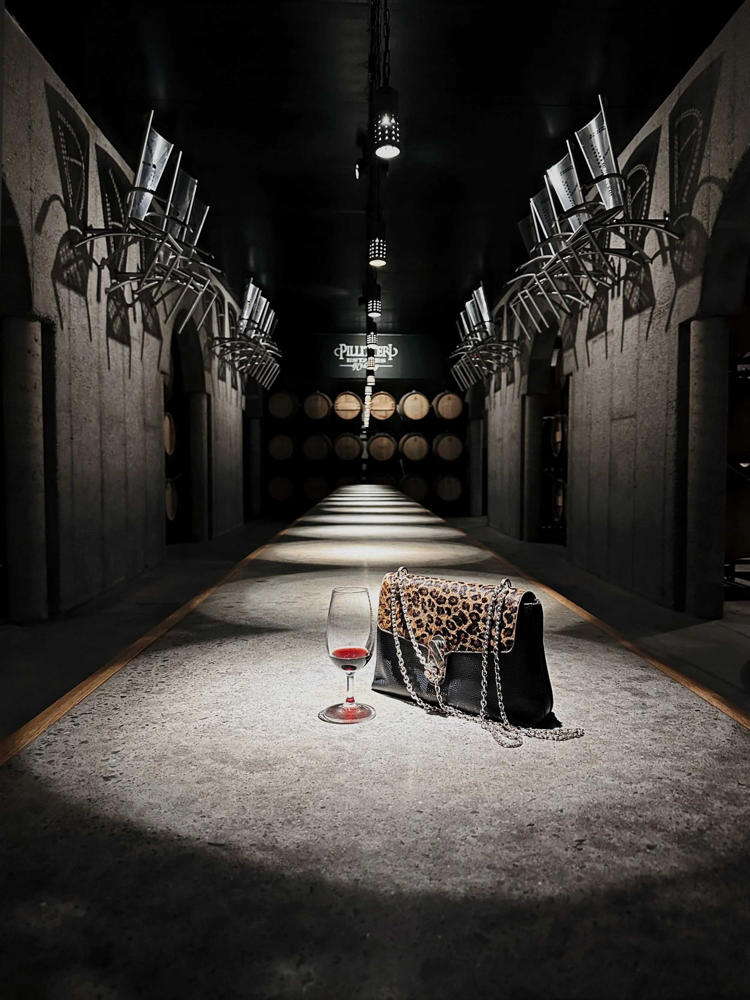A wine glass filled with red wine, a black handbag with a chain strap, and a leopard print scarf are placed on a table in a dimly lit wine cellar with barrels in the background.