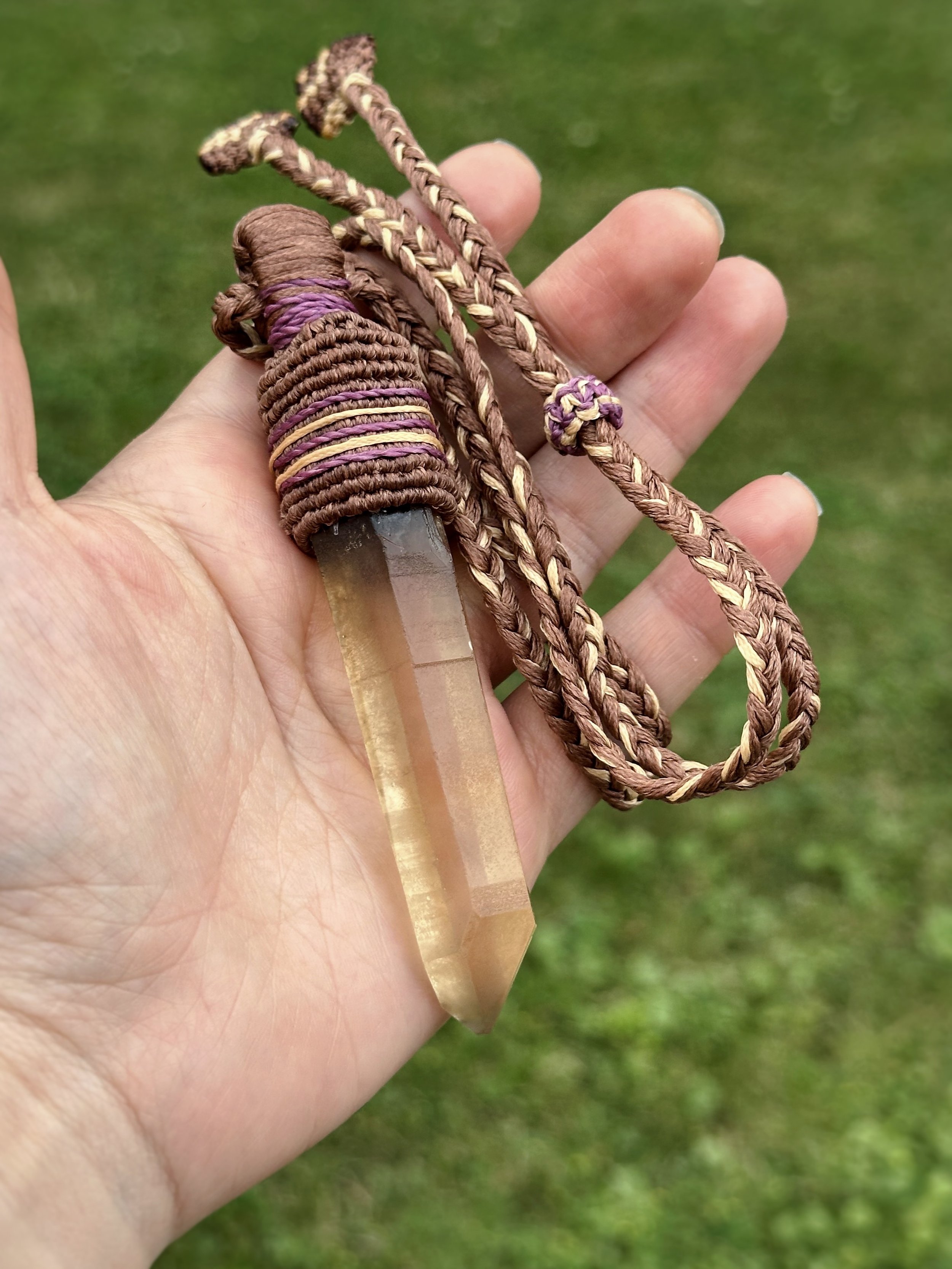 A person's hand holding a clear quartz crystal with a braided leather strap wrapped around the top, and two braided cords, against a grassy background.