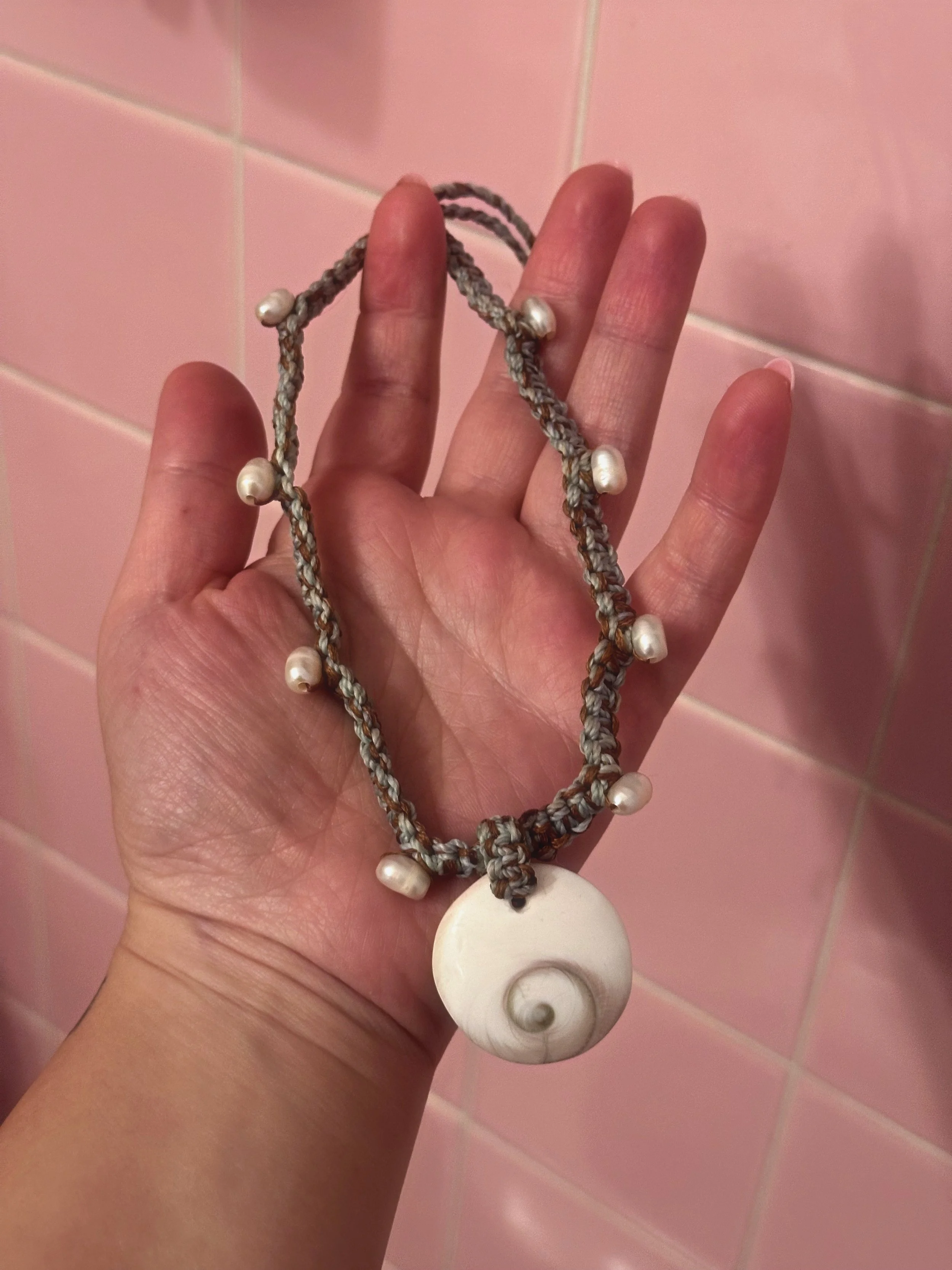A hand holding a necklace with a large spiral shell pendant and smaller shell beads, against a pink tiled wall.