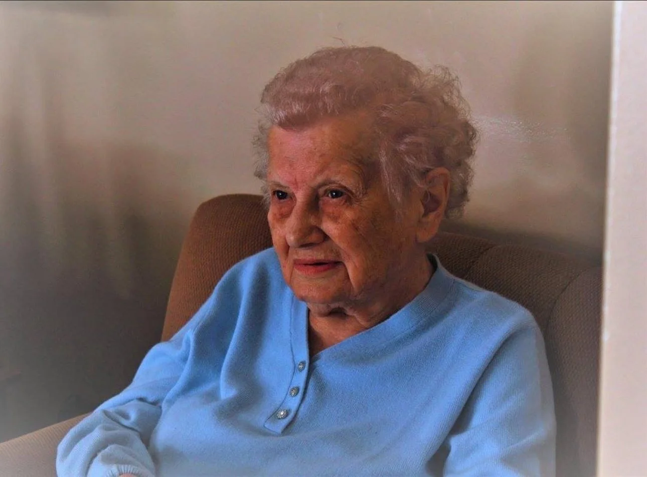 An elderly woman with curly gray hair sitting in a brown chair, wearing a light blue shirt, looking to the side inside a room with neutral-colored walls.