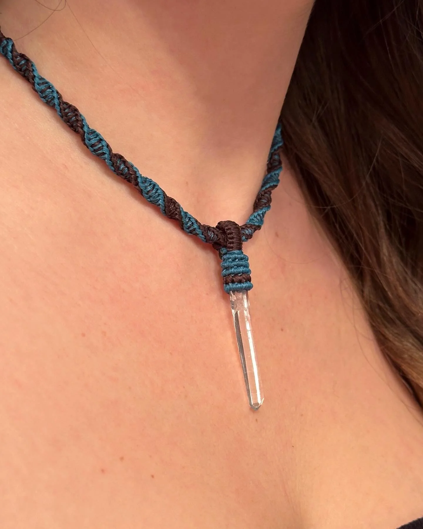 Close-up of a person wearing a handmade necklace with blue and brown braided cord and a clear quartz crystal pendant hanging from it.
