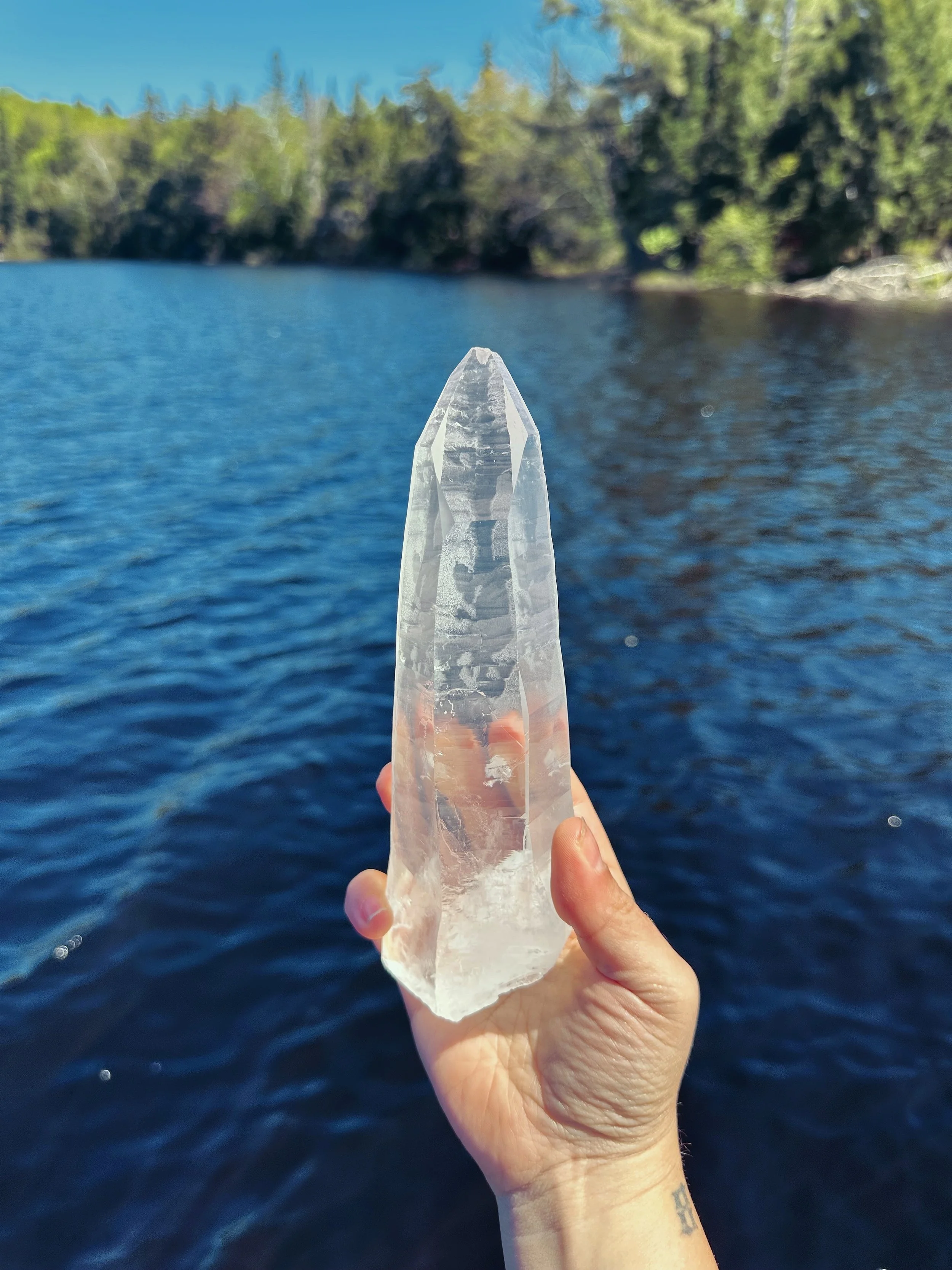 Hand holding a large, clear quartz crystal over a body of water with trees and blue sky in the background.