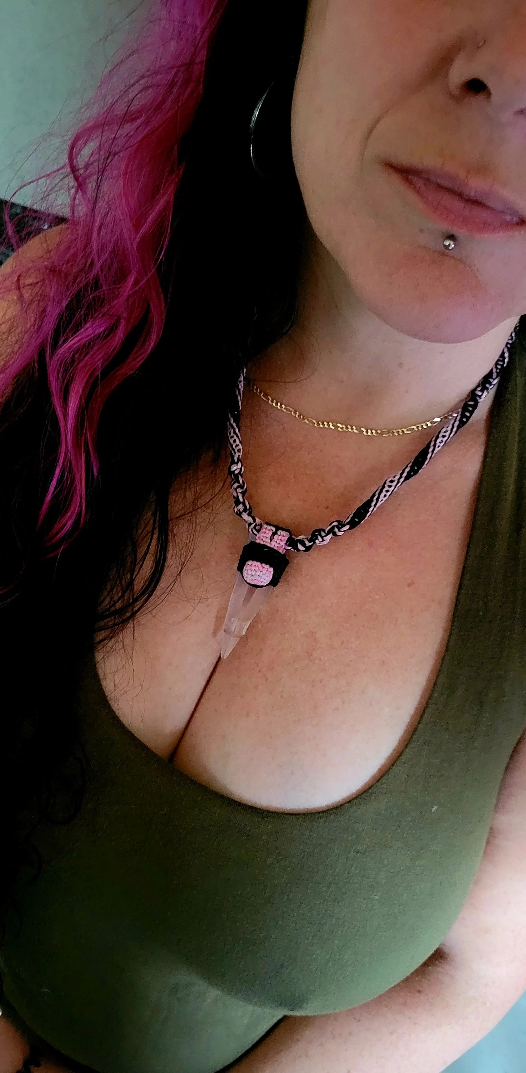 Close-up of a woman with pink and black hair, wearing a green top, gold chain necklace, and a necklace with a pendant. She has a lip piercing.